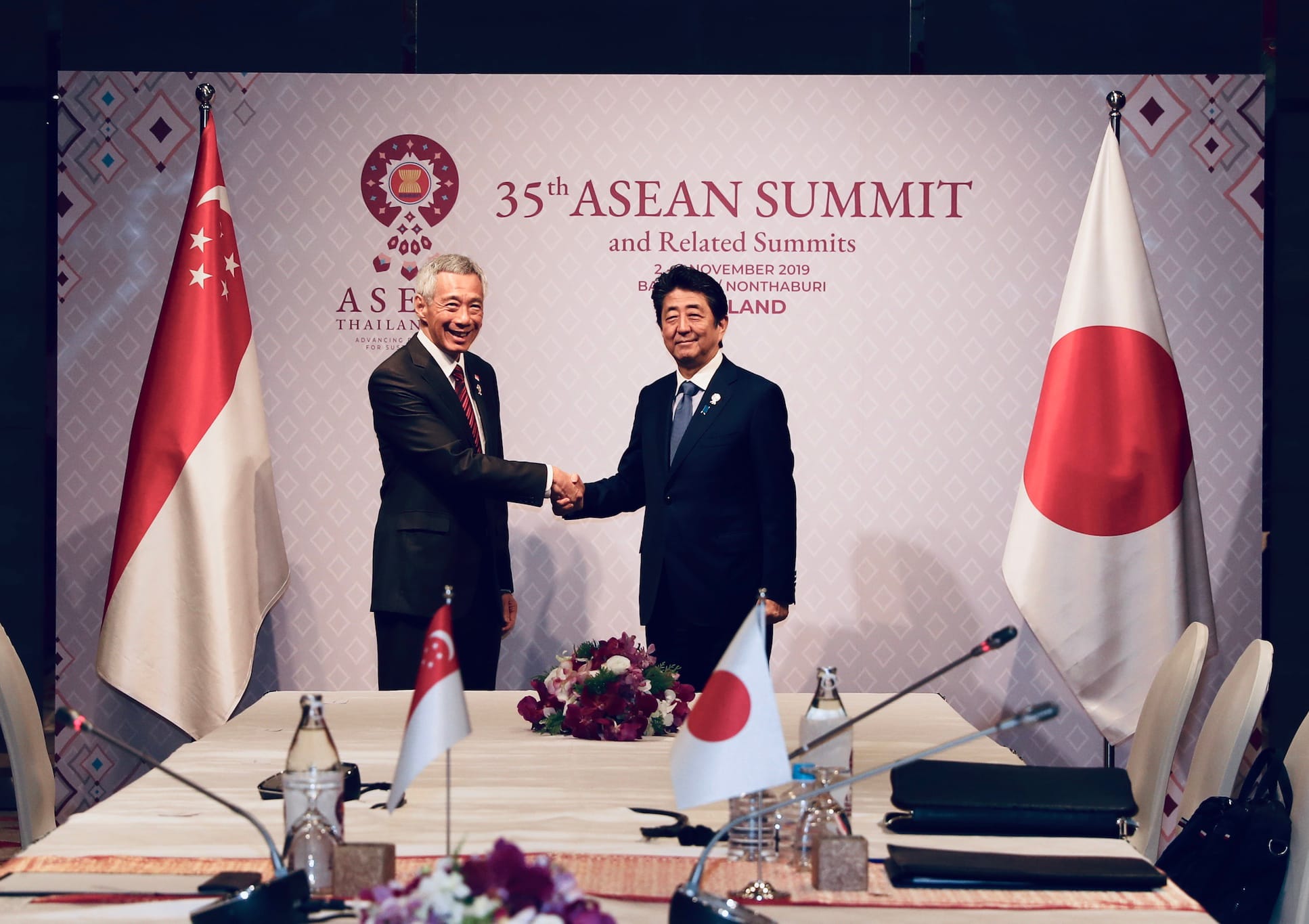 Lee Hsien Loong and Shinzo Abe shake hands at the 35th ASEAN Summit, with Singapore and Japan flags.