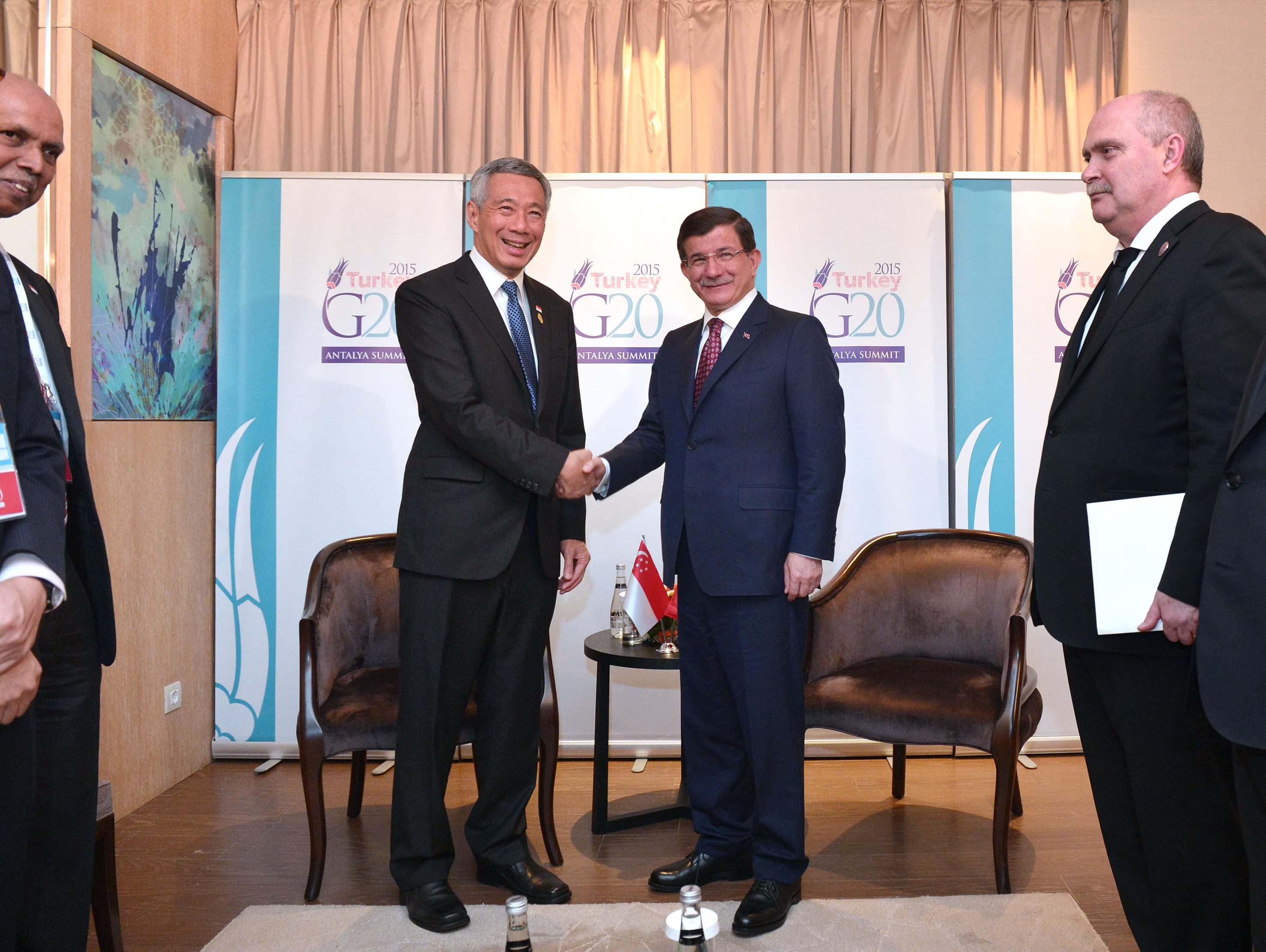 Two suited men shaking hands in front of G20 Antalya Summit backdrop. Others stand by.