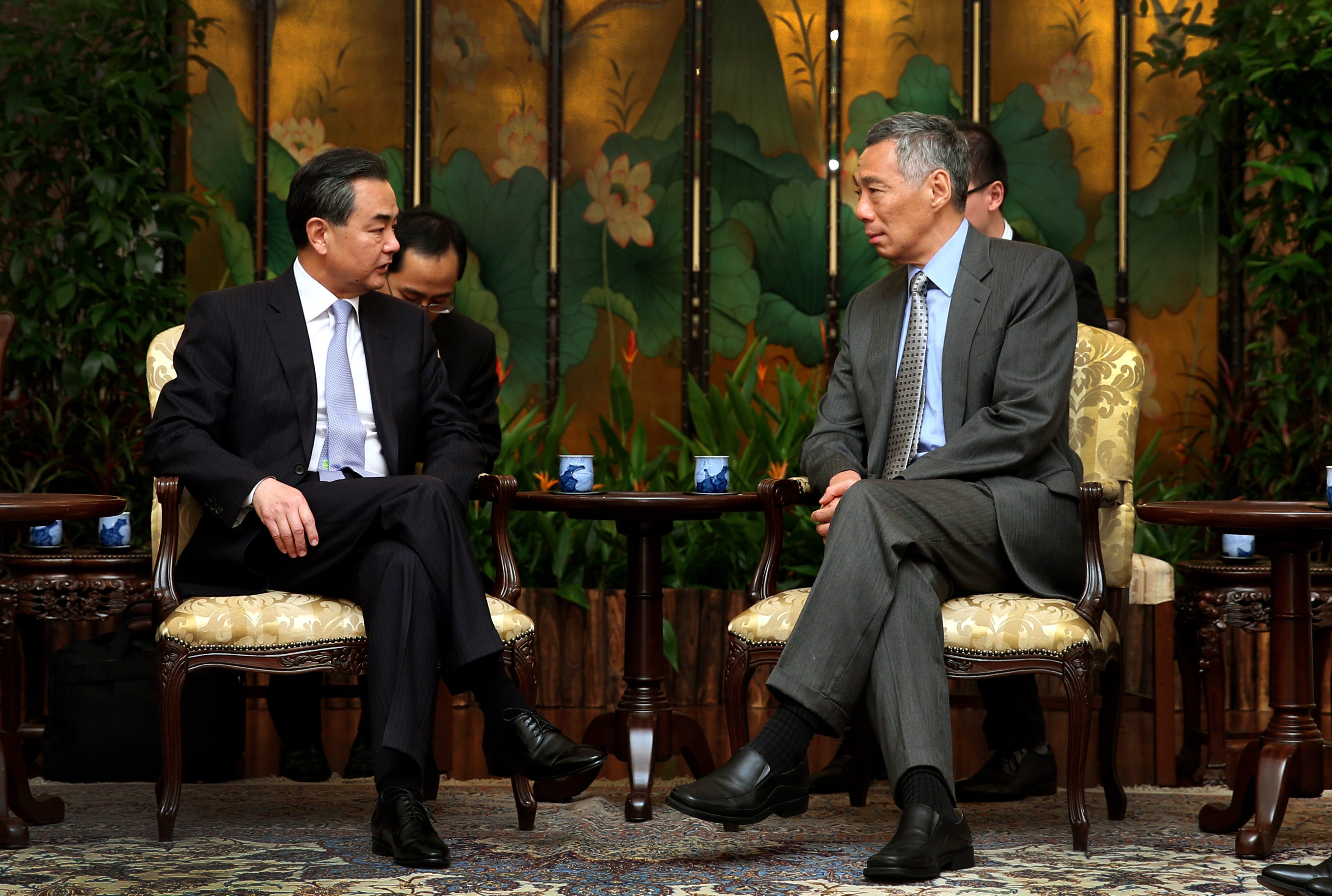 Two men in suits seated, facing each other against a floral screen backdrop.
