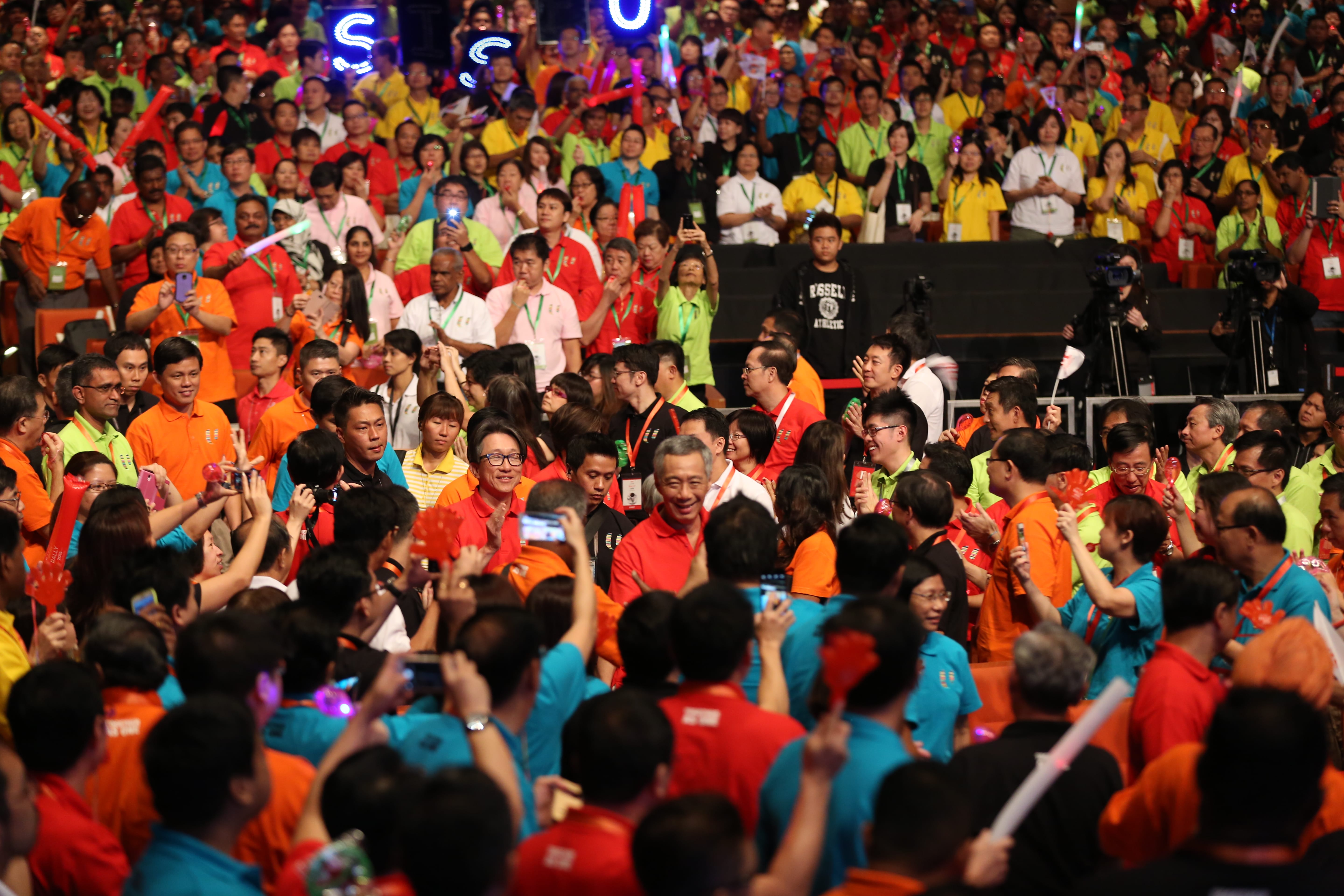 Large crowd of people in colorful shirts holding up phones and cheering.