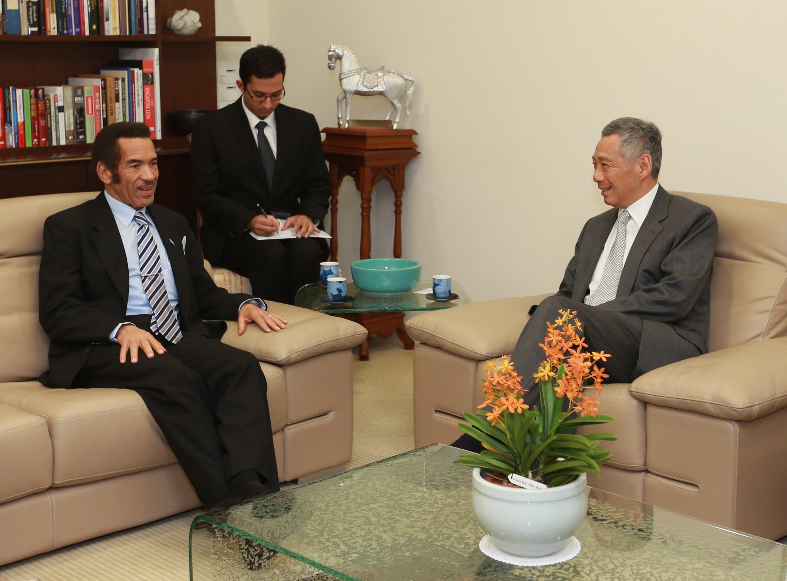 Two men in suits seated on couches, another taking notes. Bookshelf, horse statue visible.