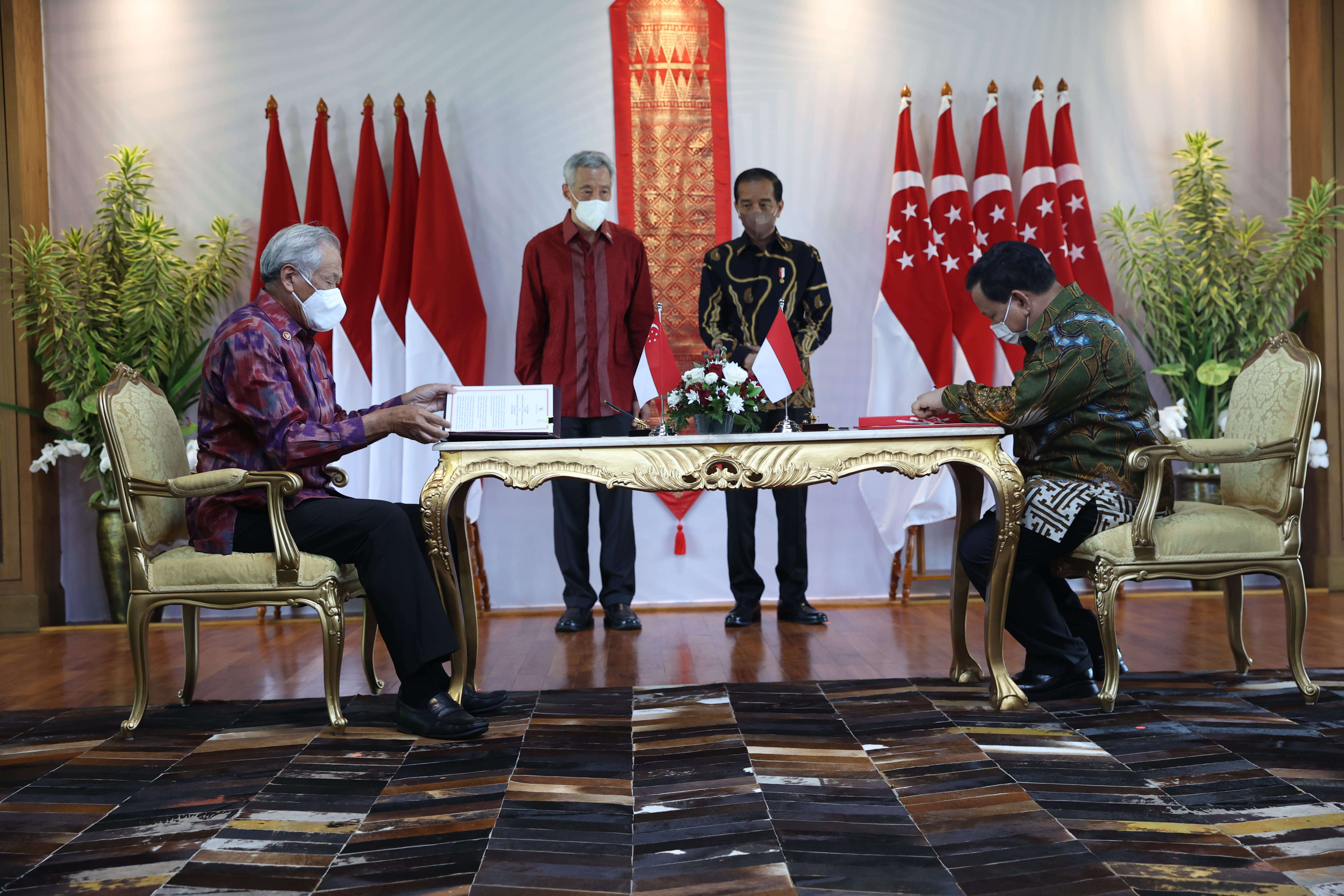 Two seated figures sign document at table flanked by Indonesian & Singaporean flags and Lee Hsien Loong and Joko Widodo.