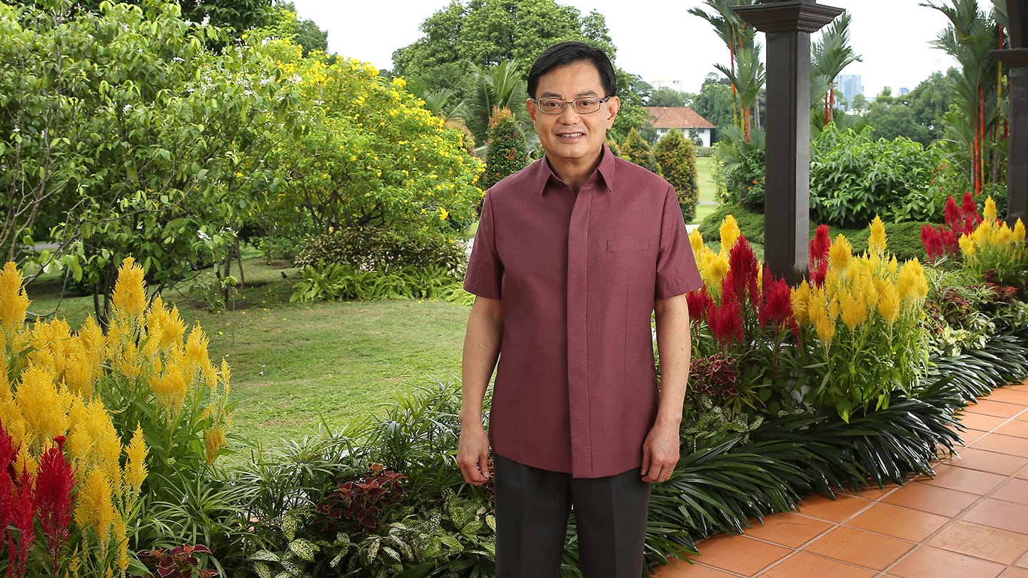 Man in glasses and maroon shirt stands amongst red and yellow flowers in a garden.