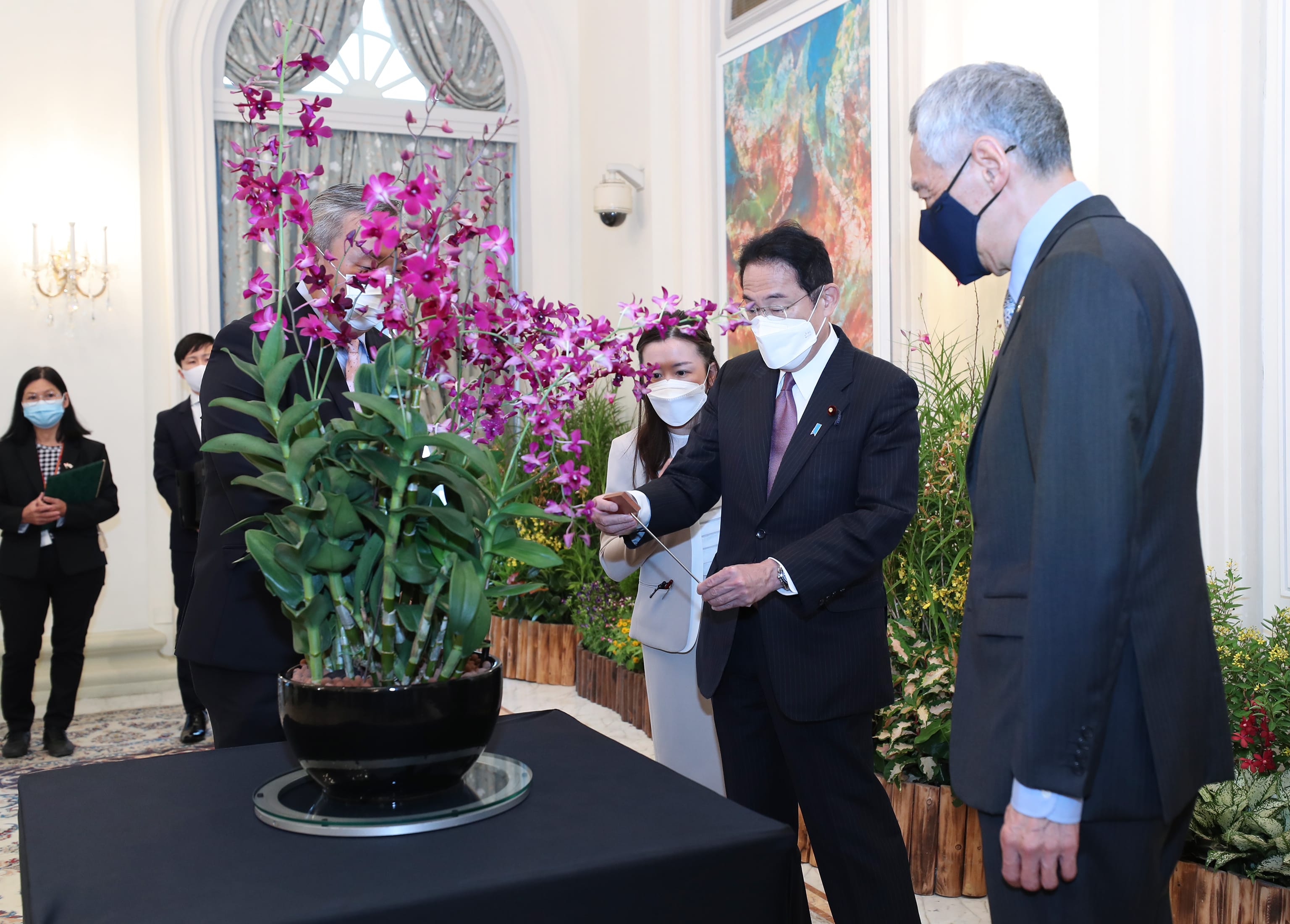 Fumio Kishida and Lee Hsien Loong, both masked, stand with purple orchids in a dark pot.