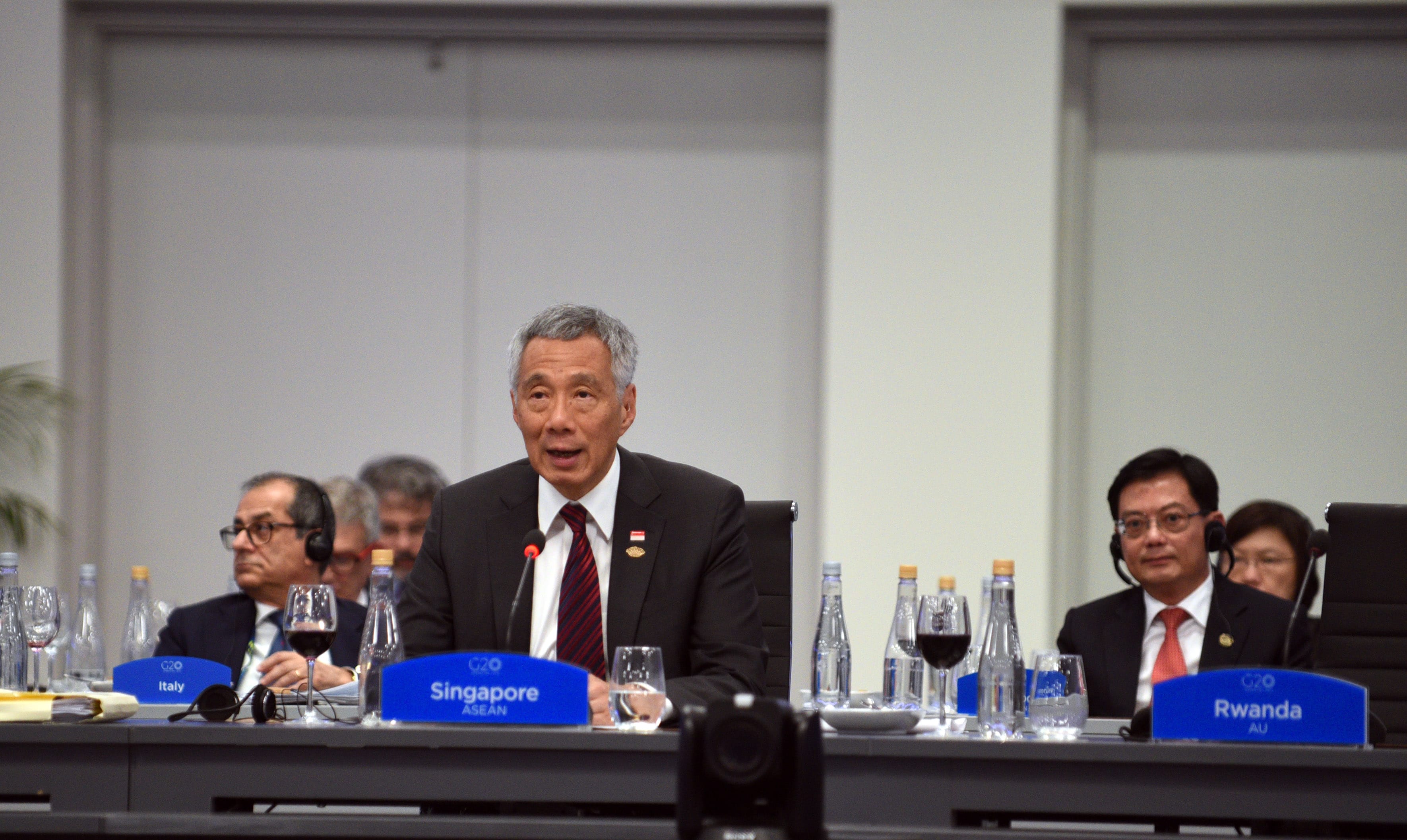 Lee Hsien Loong speaking at G20 meeting, with "Singapore" nameplate visible. Delegates at table with water, wine.