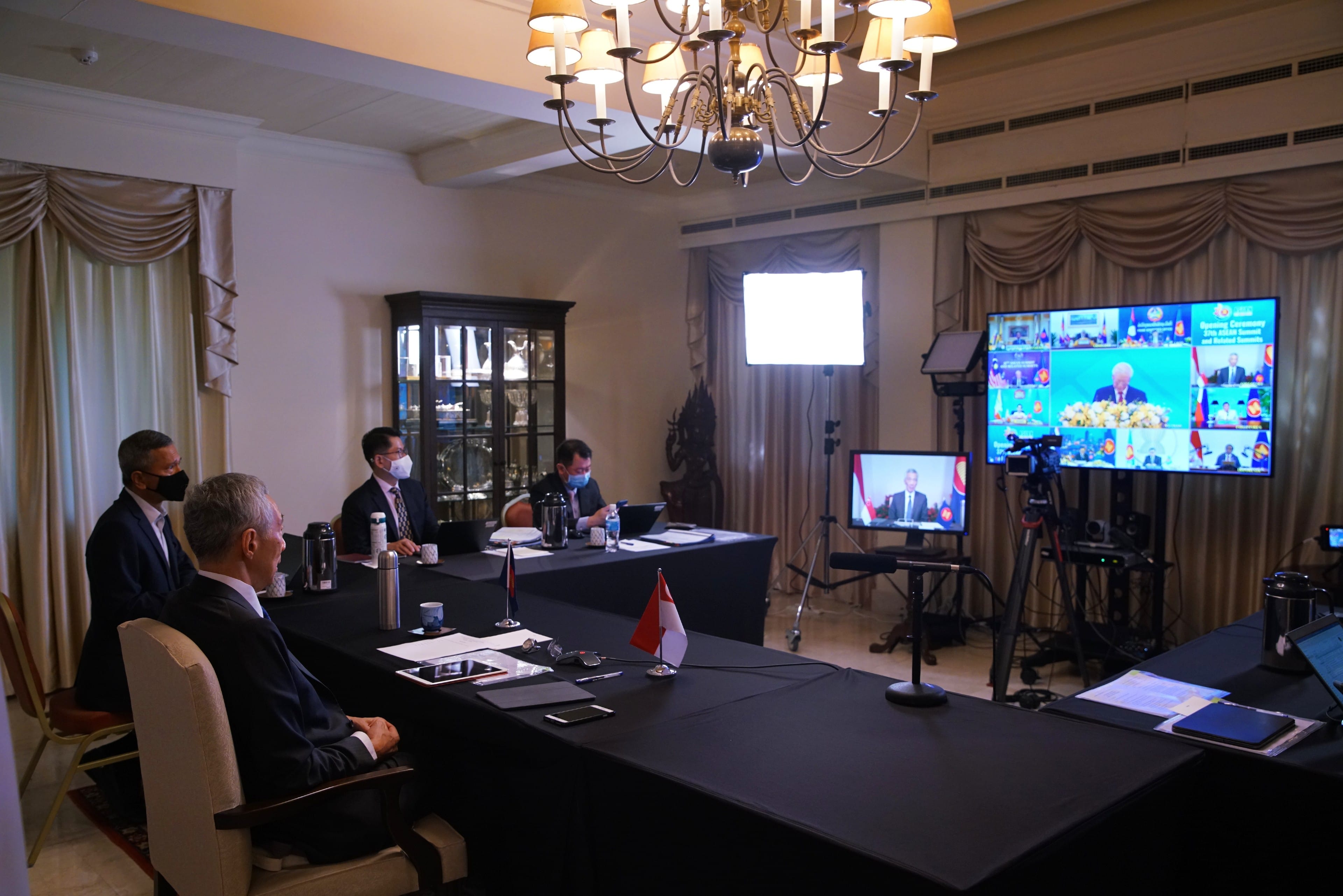 People in suits at long tables with masks, live screens with broadcast; chandelier overhead.