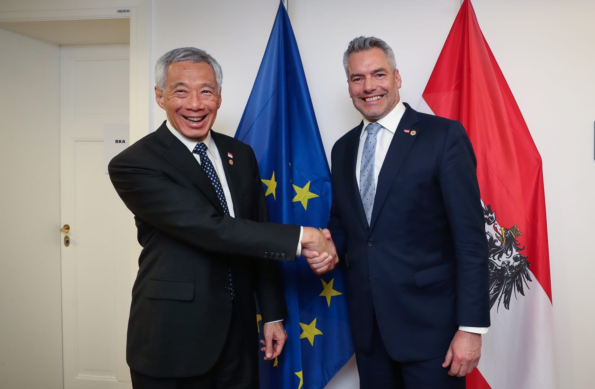 Two men in suits shake hands, with EU and Austria flags in the background.