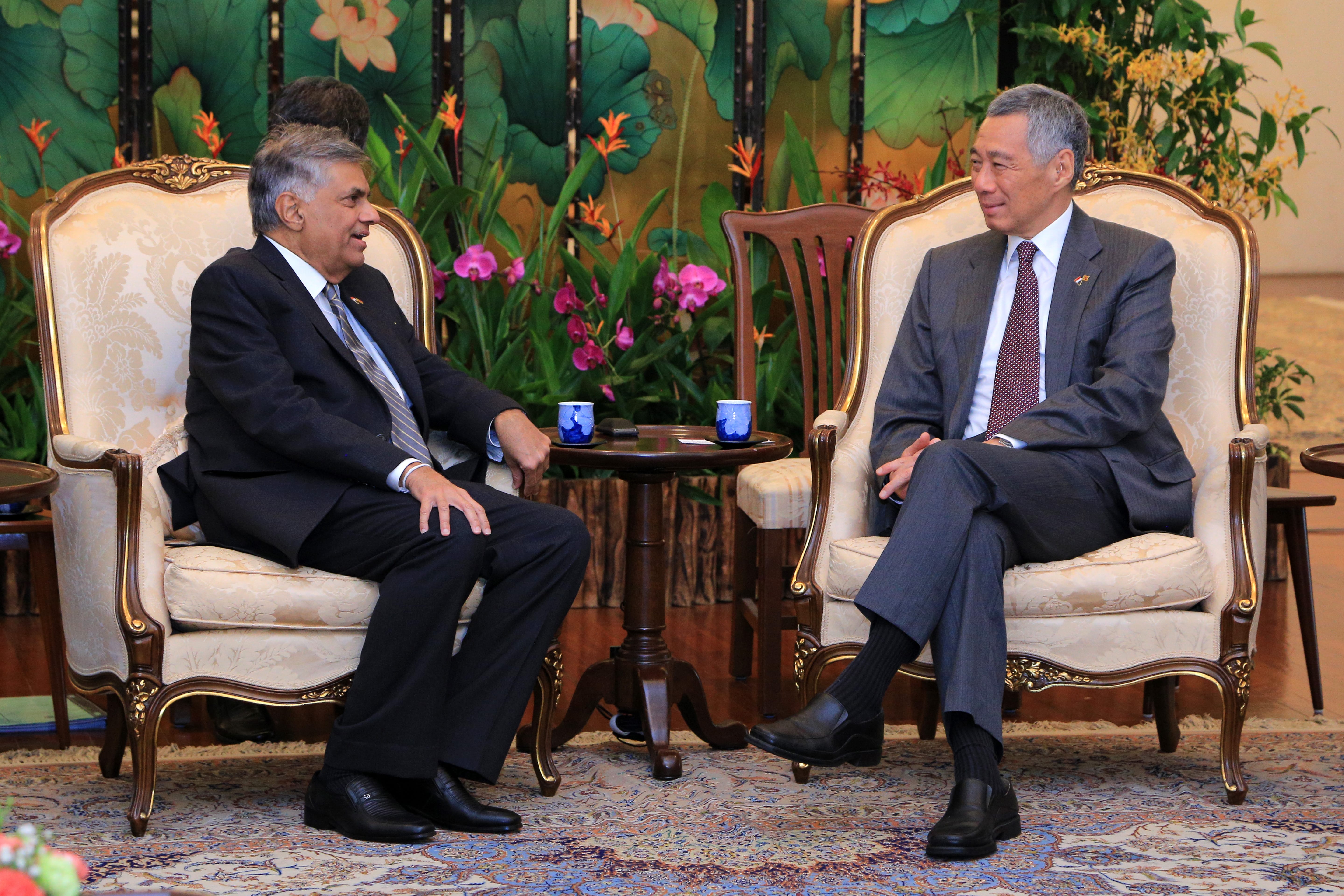 Two men in suits seated in ornate chairs facing each other with a floral backdrop.