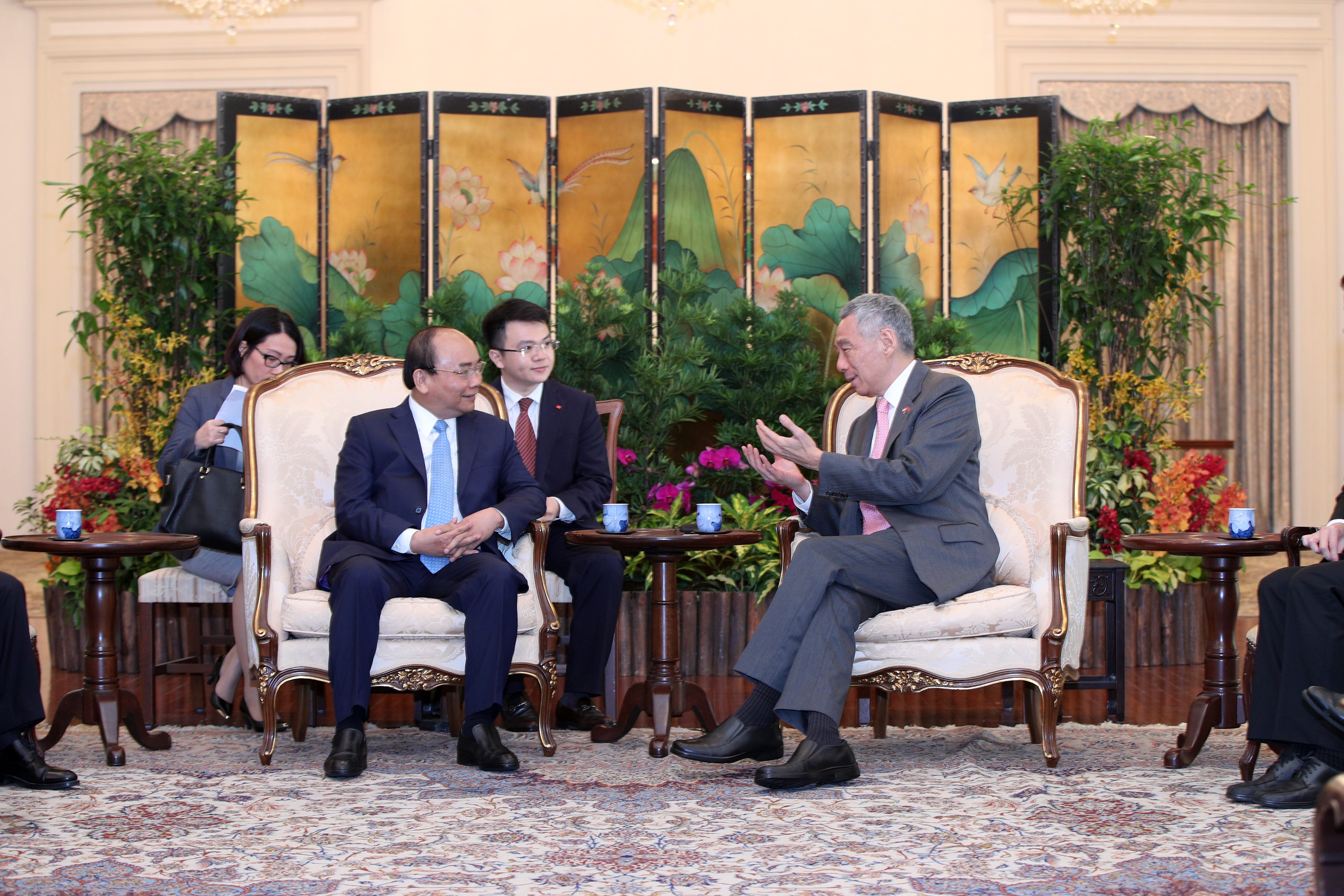 Two men in suits sitting on ornate chairs, gesturing during a meeting, with a floral screen behind them.