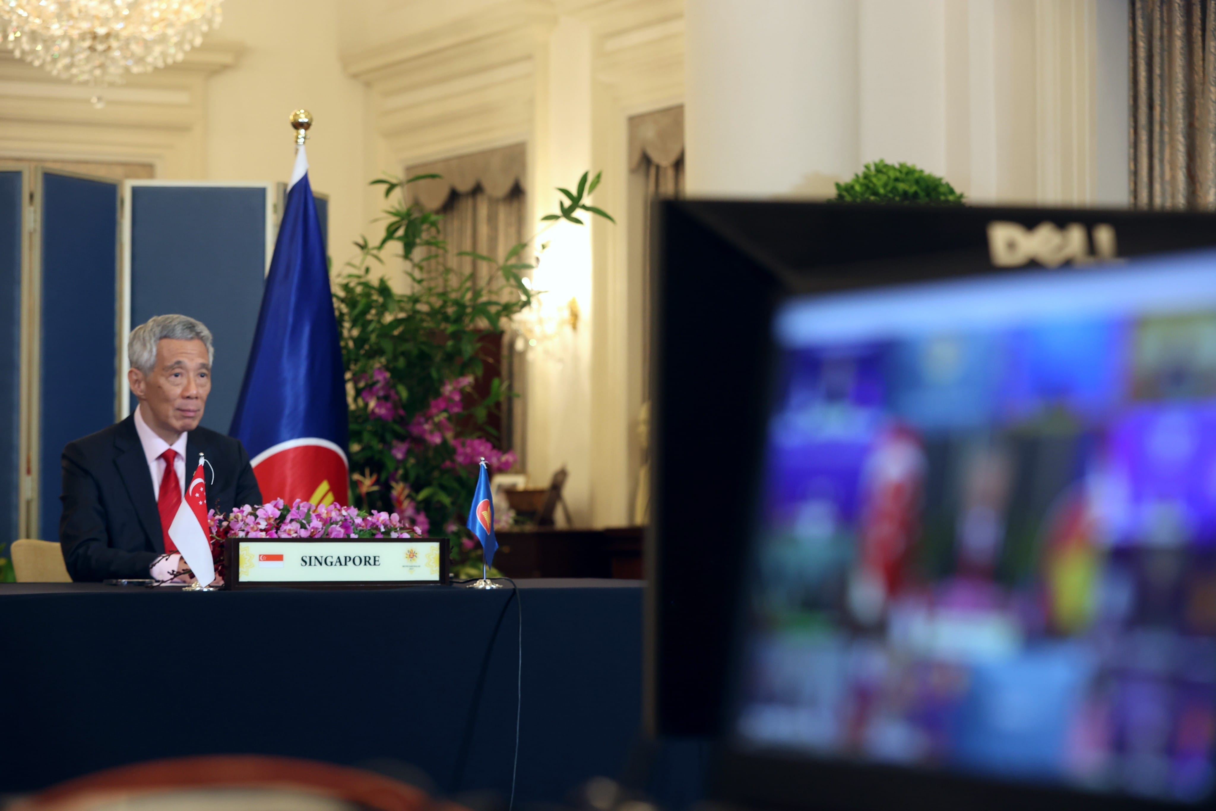 Lee Hsien Loong at table with Singapore flag, ASEAN flag, and Dell monitor showing video conference.