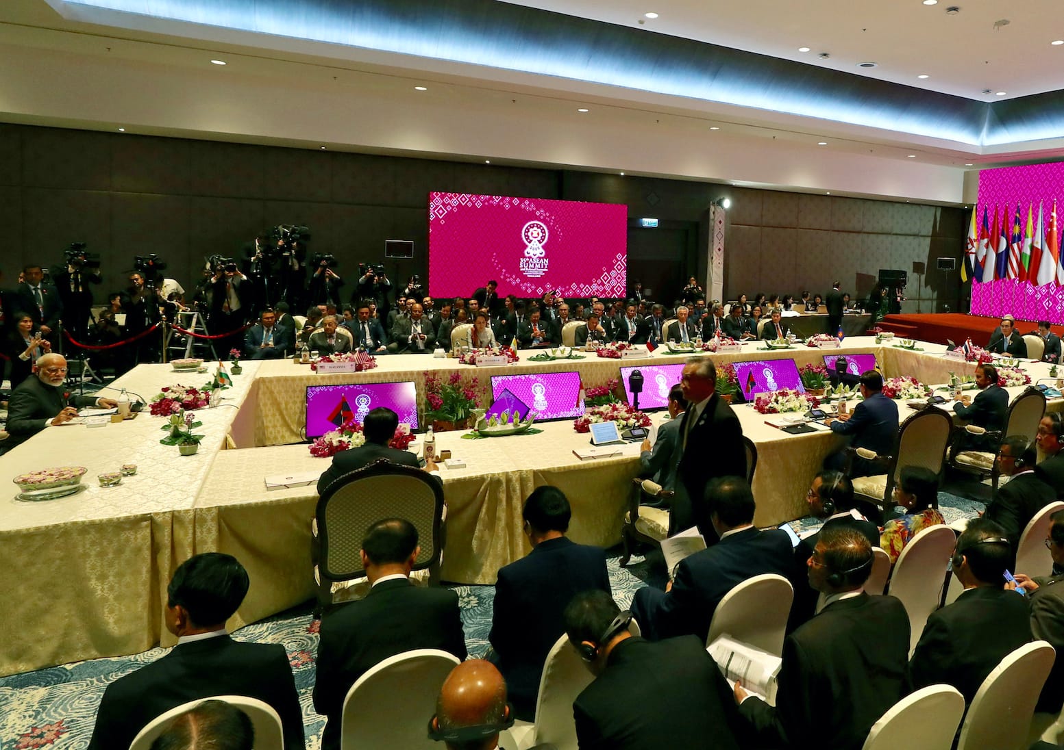 ASEAN summit meeting with attendees at a table, press at the back, flags and screens visible.