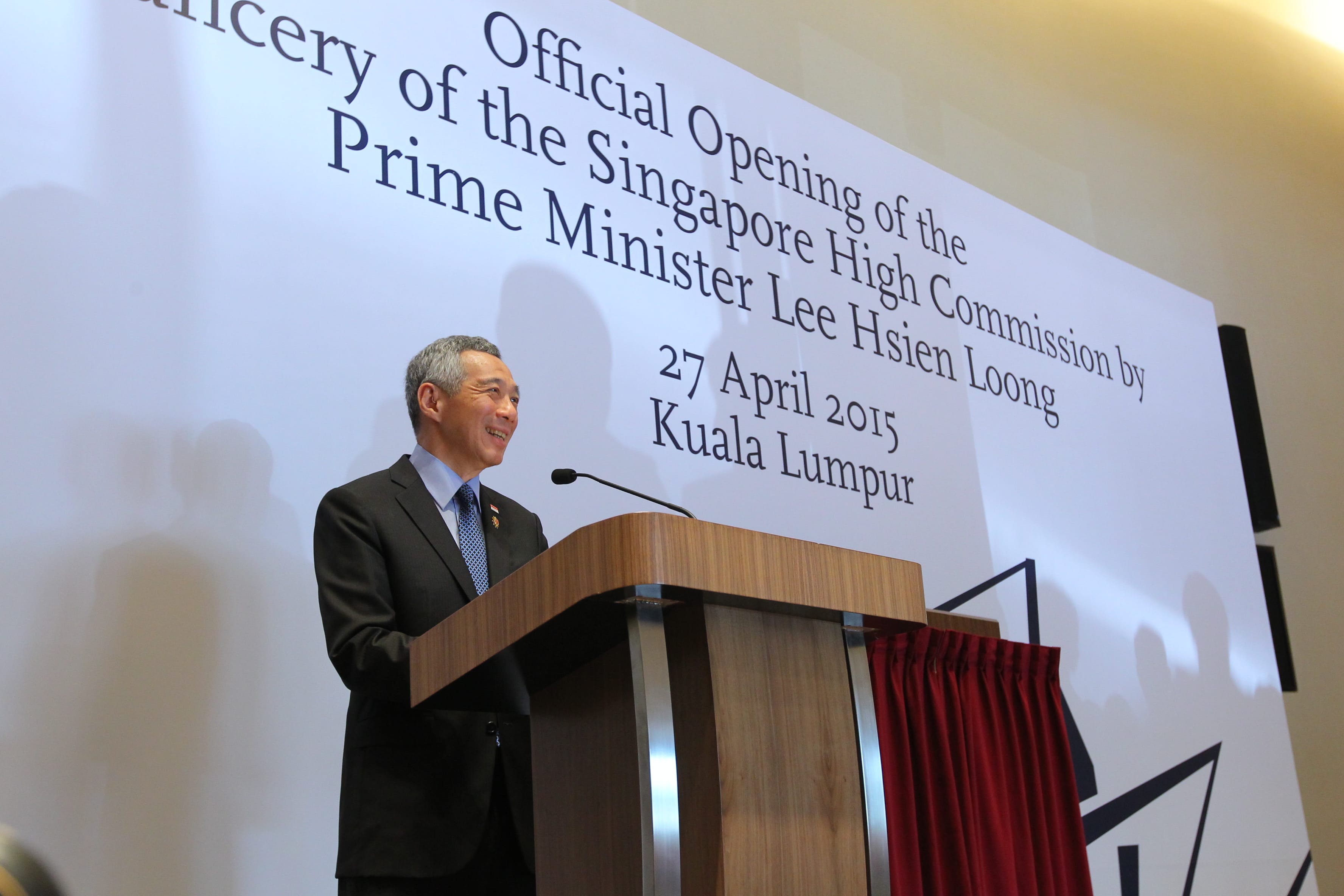 Lee Hsien Loong at podium in suit, giving a speech. Backdrop reads: "Official Opening...".