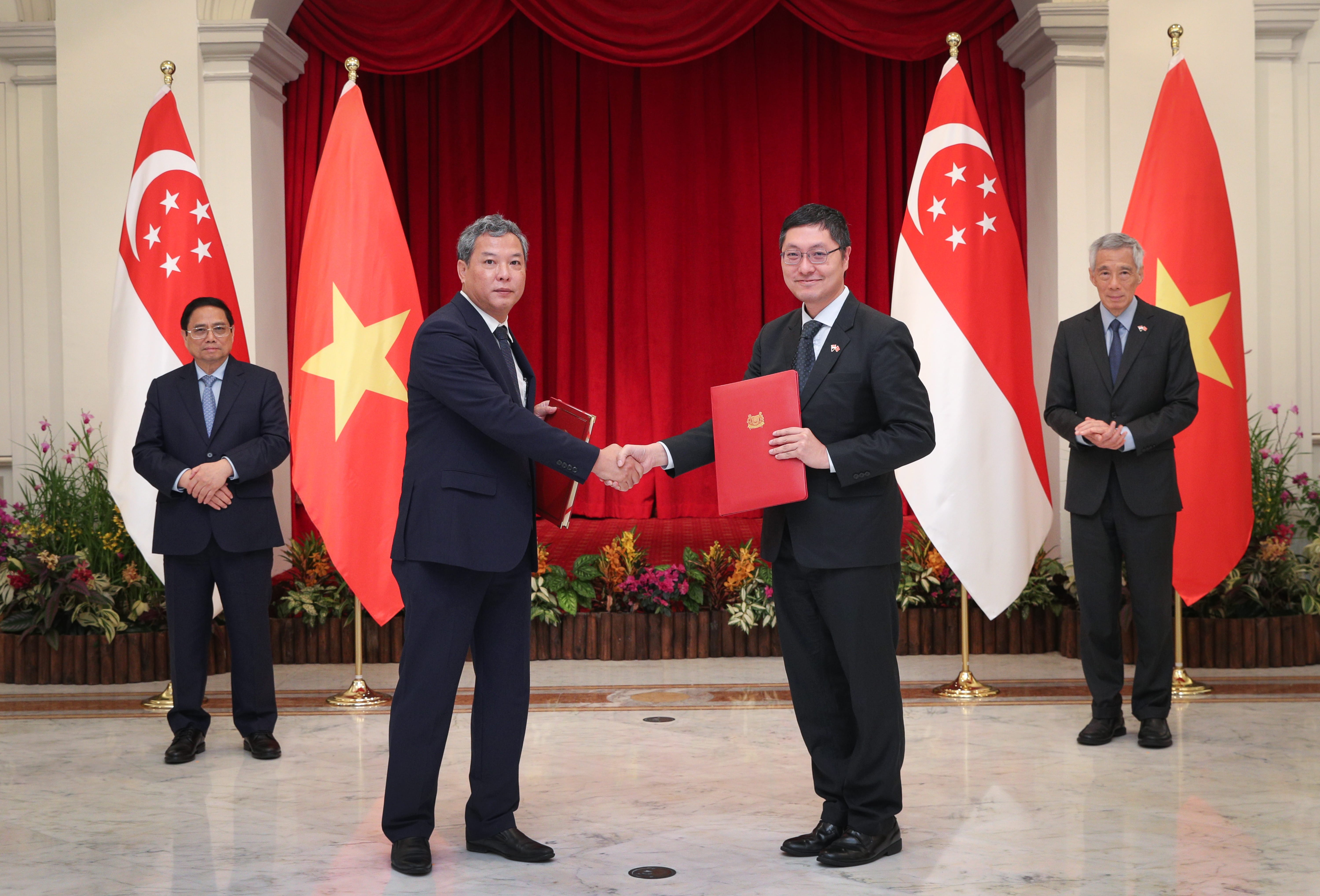 Two men in suits shake hands, one holding a red folder, with Singapore & Vietnam flags behind them. Two other men stand nearby.