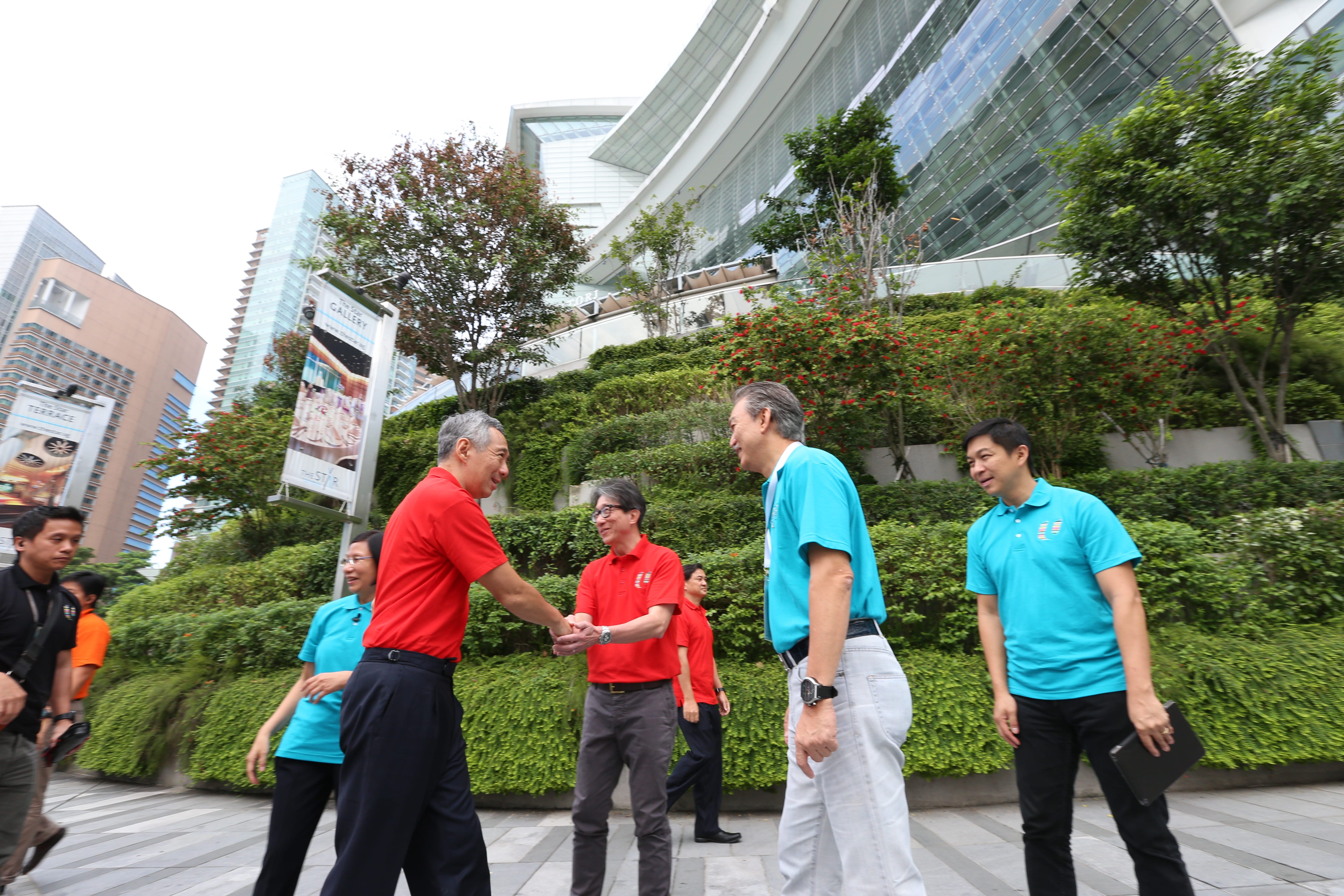 Lee Hsien Loong in red shirt shaking hands, outdoors near a building with glass facade and greenery.