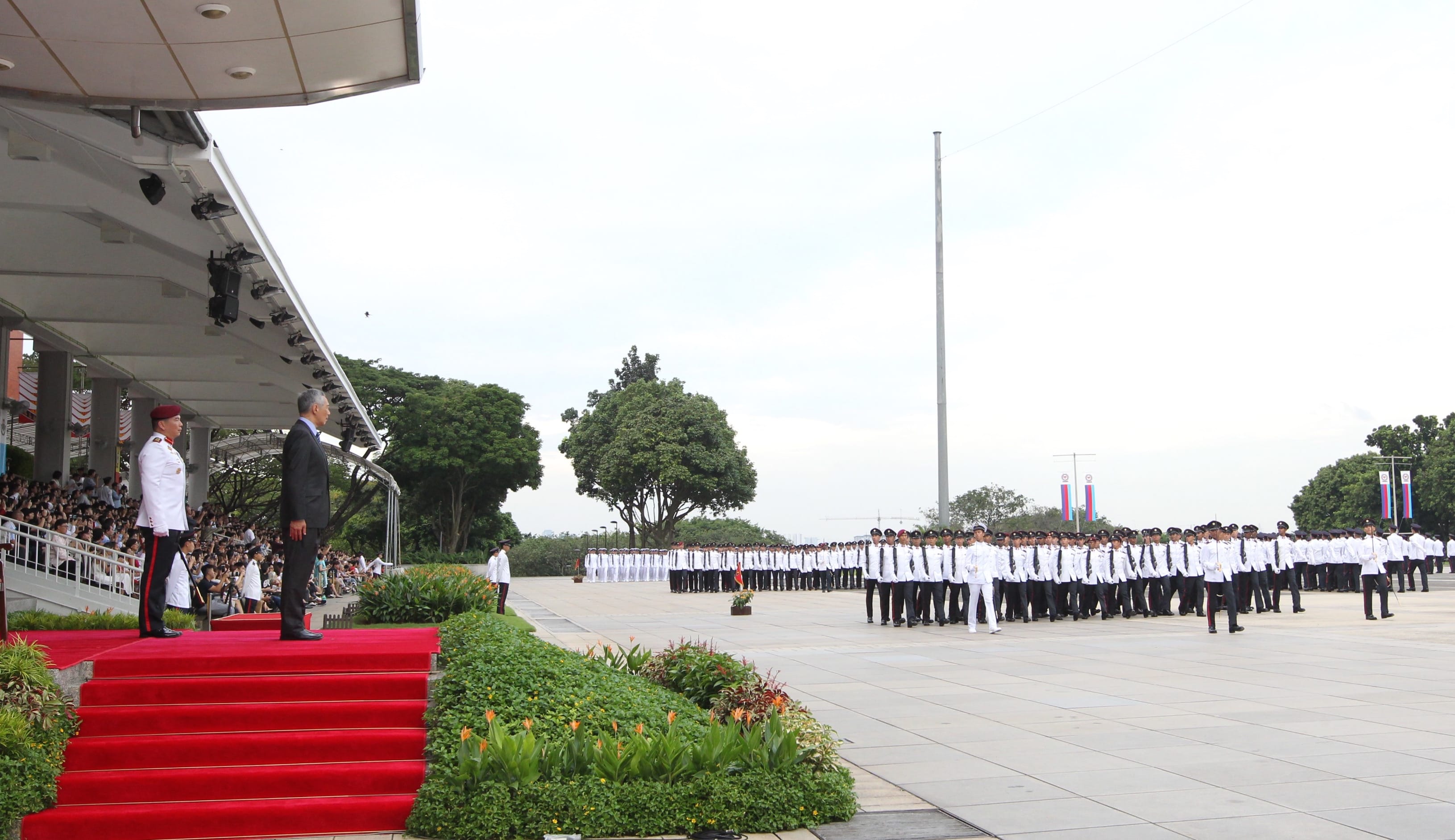 Lee Hsien Loong, in suit, and guard stand on red carpeted steps. Military parade stands at attention in background.