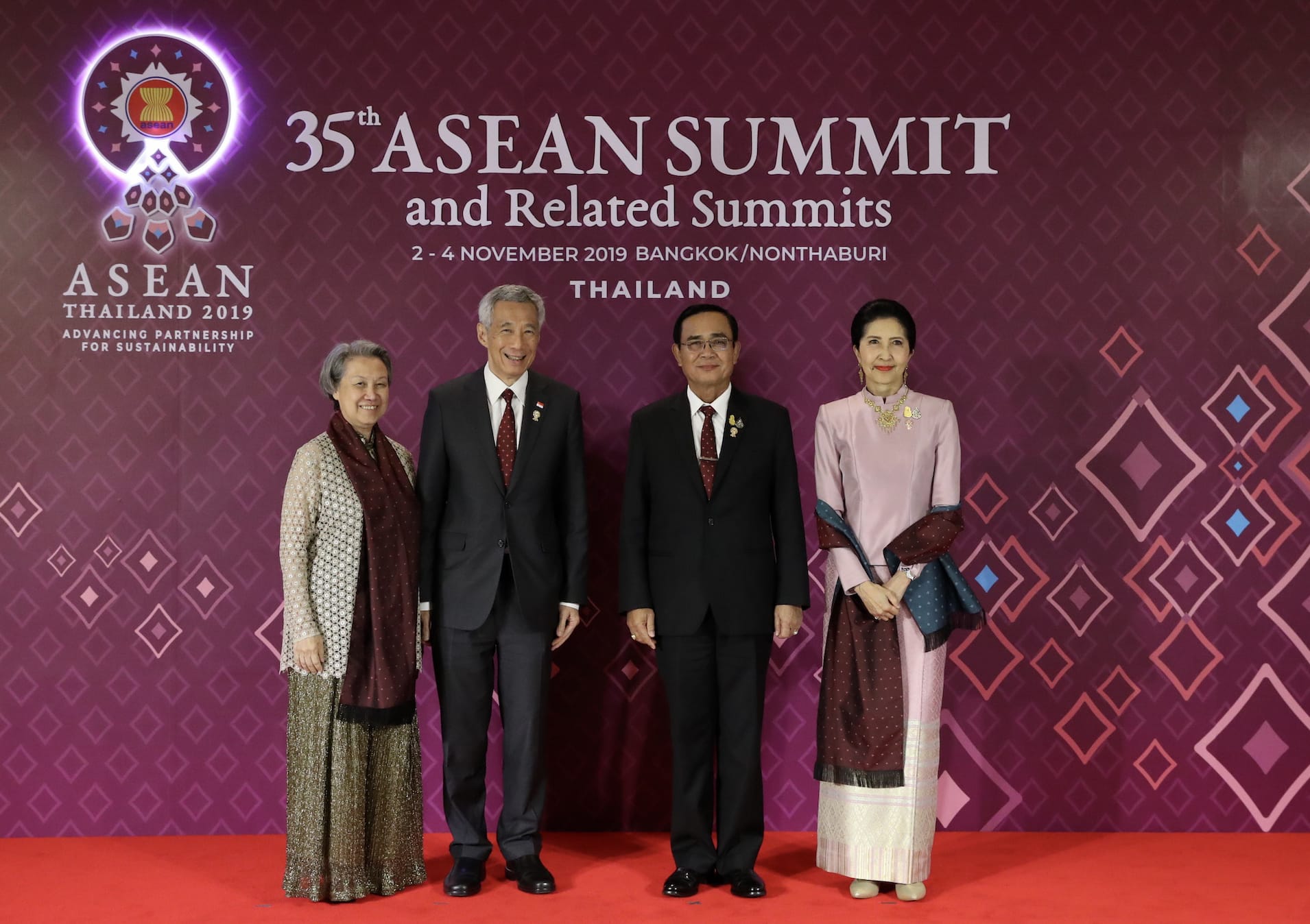 Four people stand before a "35th ASEAN Summit" backdrop. Two men in suits flank two women in traditional attire.