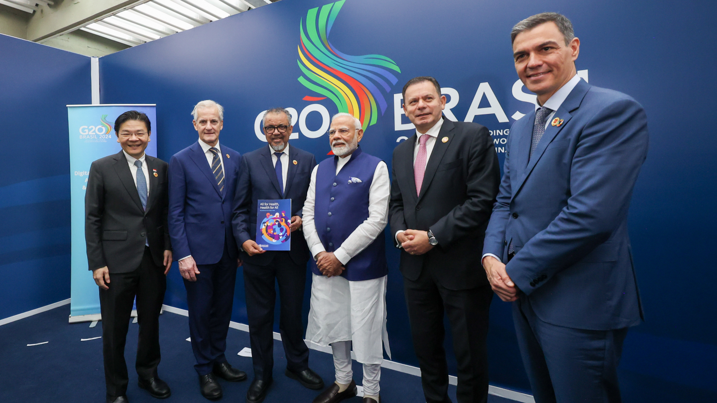 Six people in suits stand before a G20 Brazil 2024 backdrop. One holds a booklet titled "All for Health, Health for All."