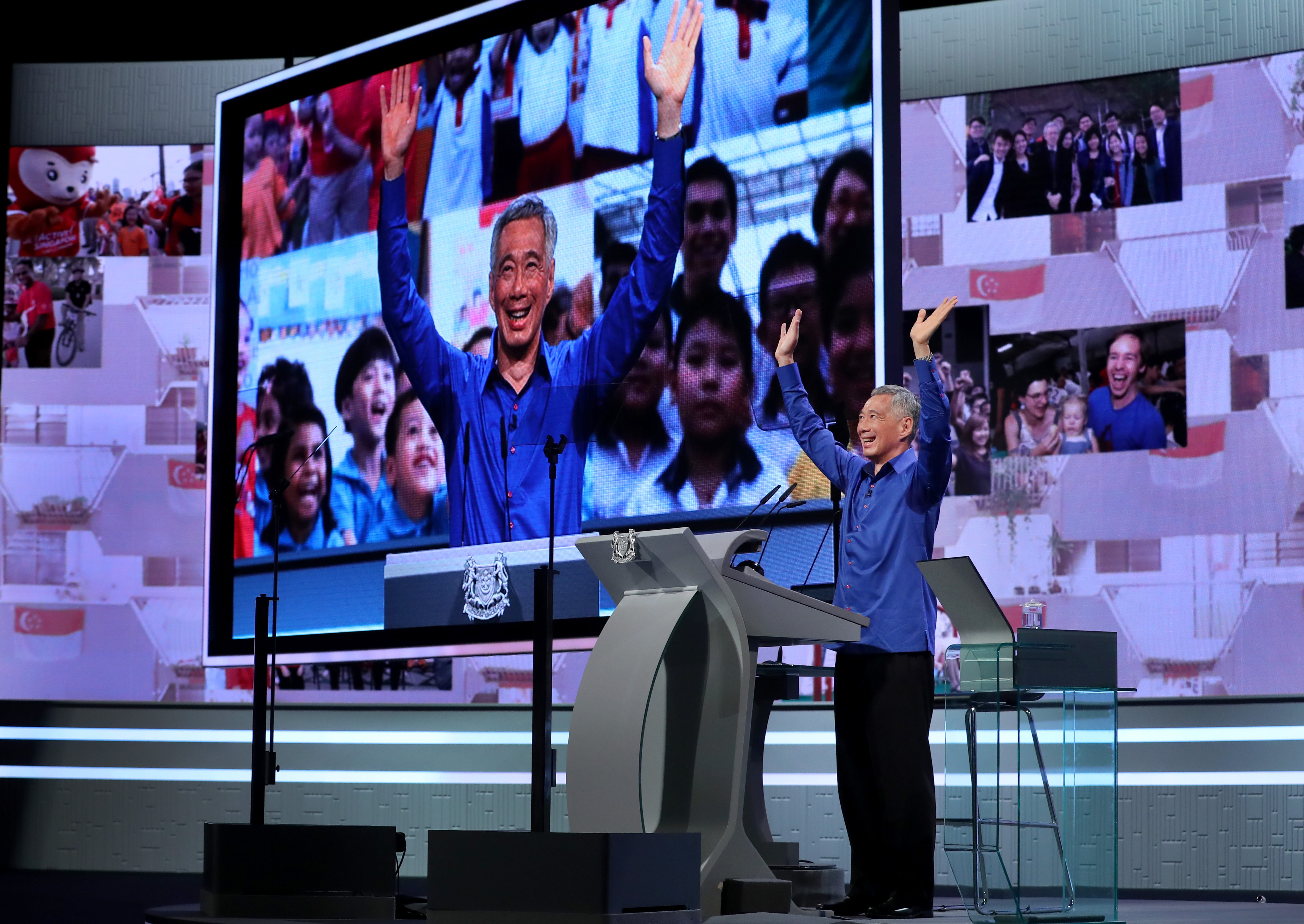 Lee Hsien Loong raises hands behind a podium, a screen behind shows his image among others raising hands.