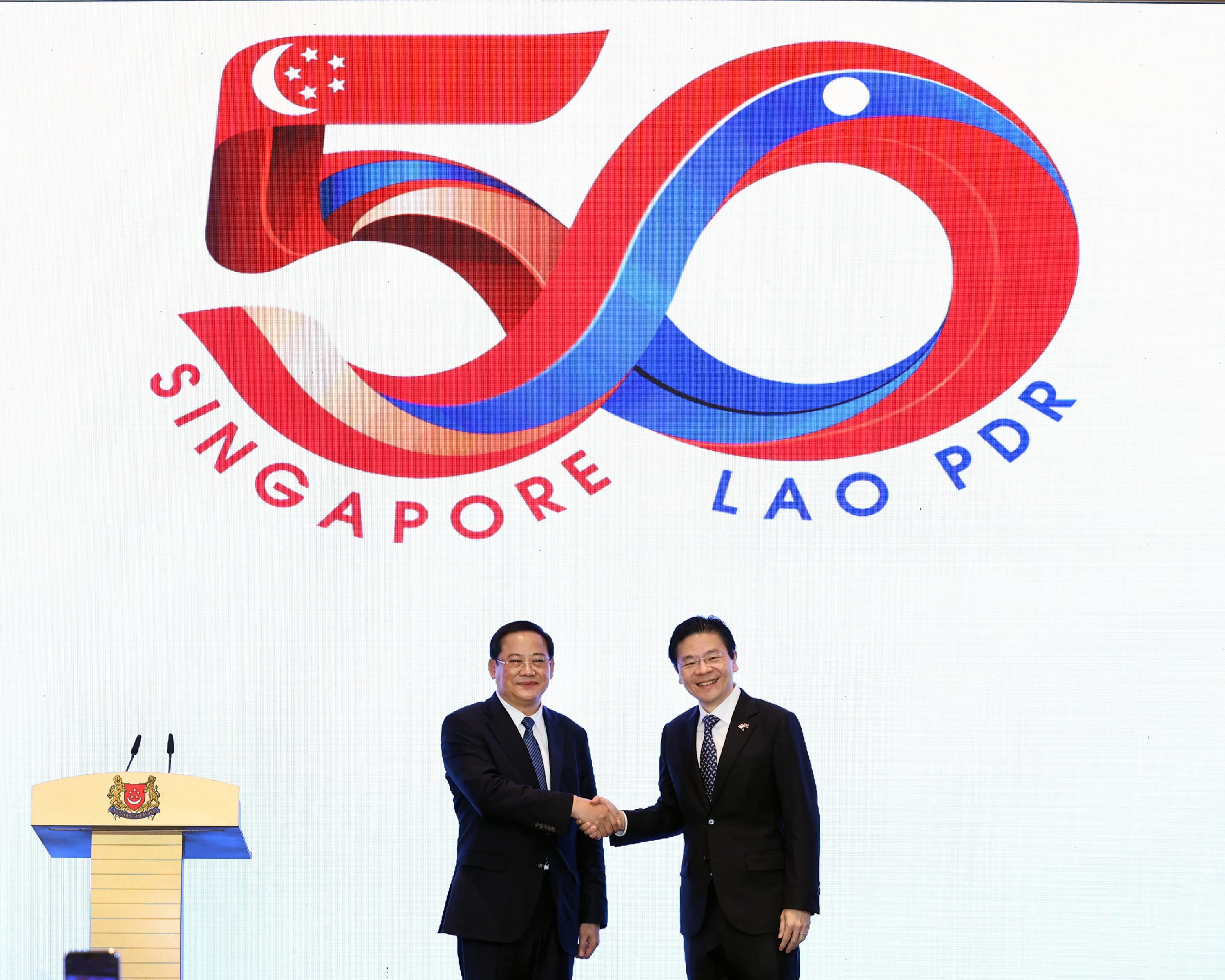 Two men in suits shake hands before a "50 SINGAPORE LAO PDR" graphic. Podium on left with Singapore coat of arms.