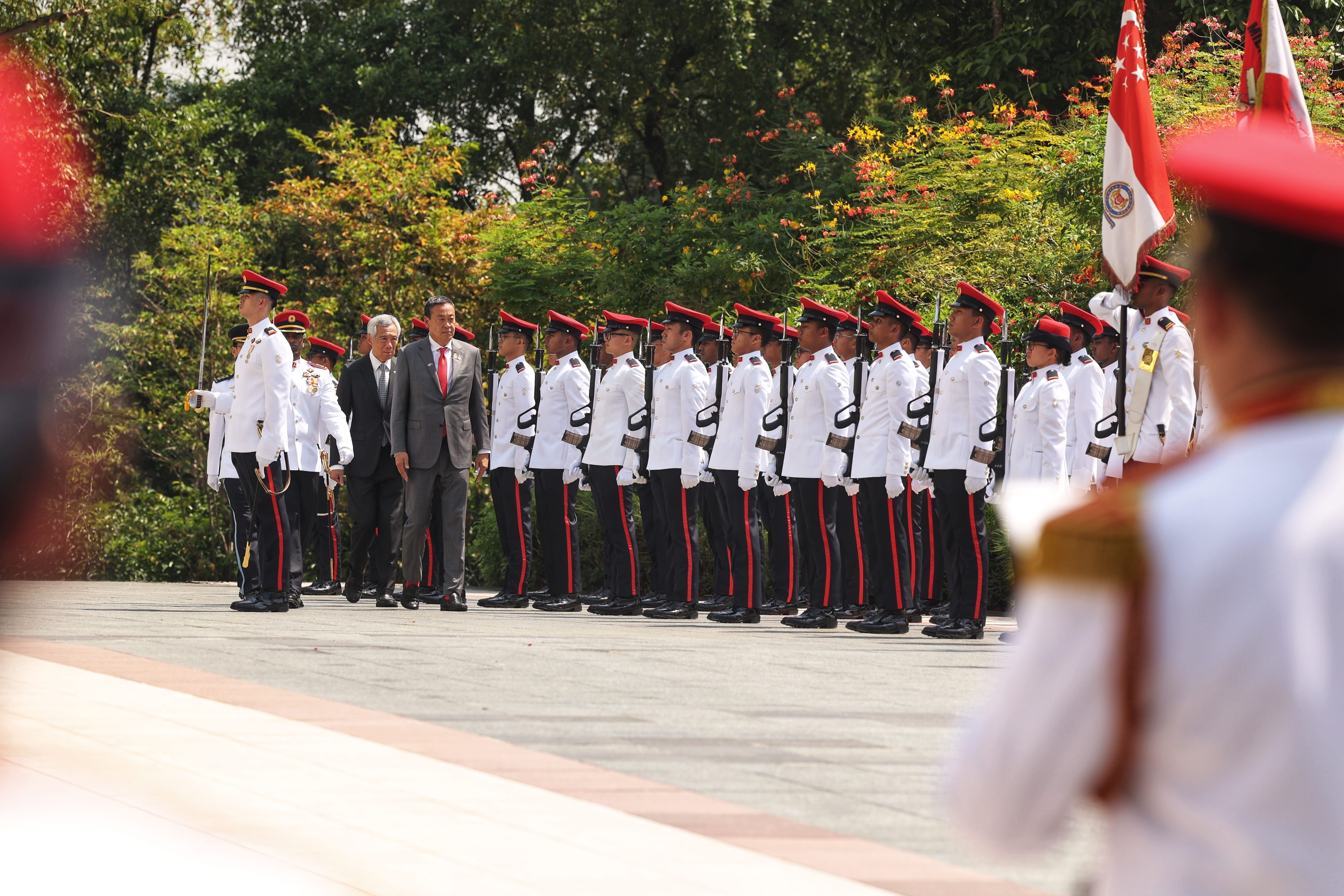 Two men in suits walking alongside a line of soldiers in white uniforms and red caps.