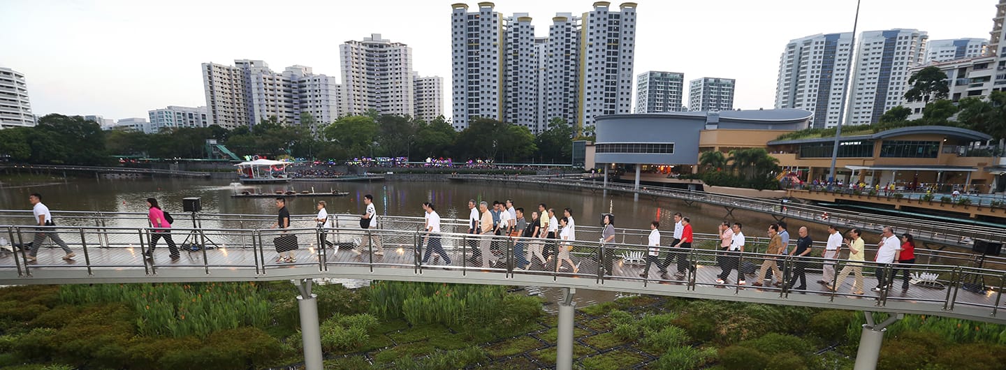 People walk on a bridge over water; city buildings in background.
