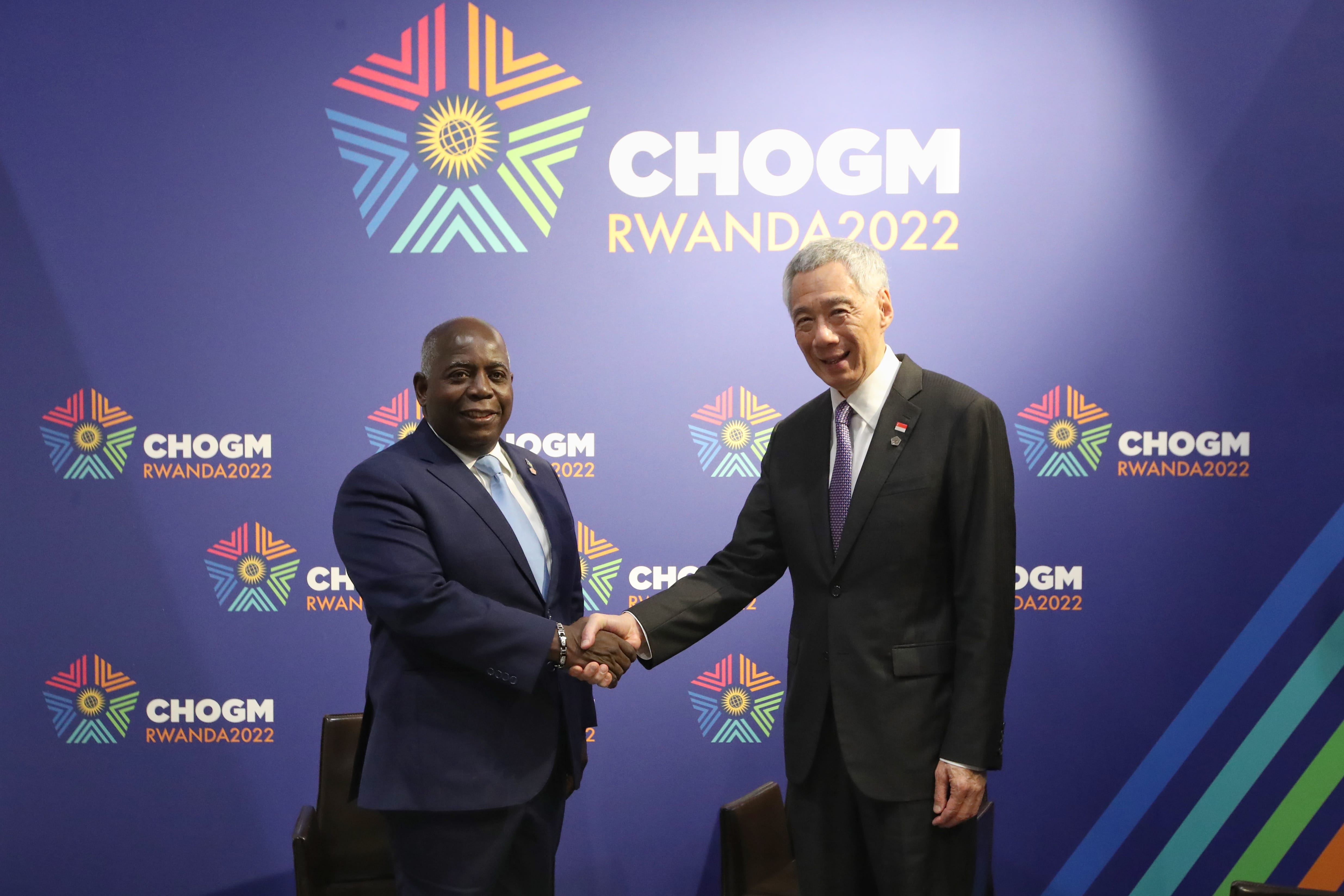 Two men in suits shake hands in front of a CHOGM RWANDA2022 backdrop.