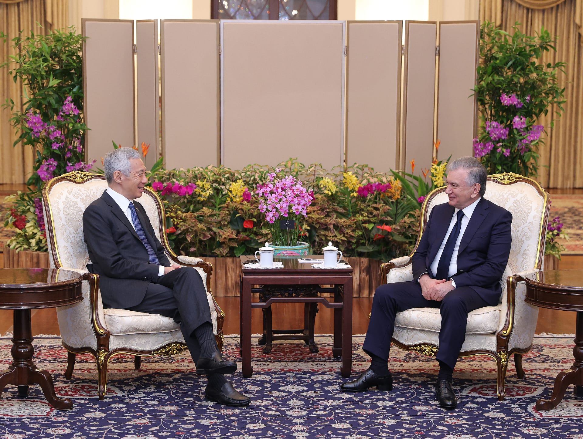 Two men in suits seated in ornate chairs face each other, flowers between them.