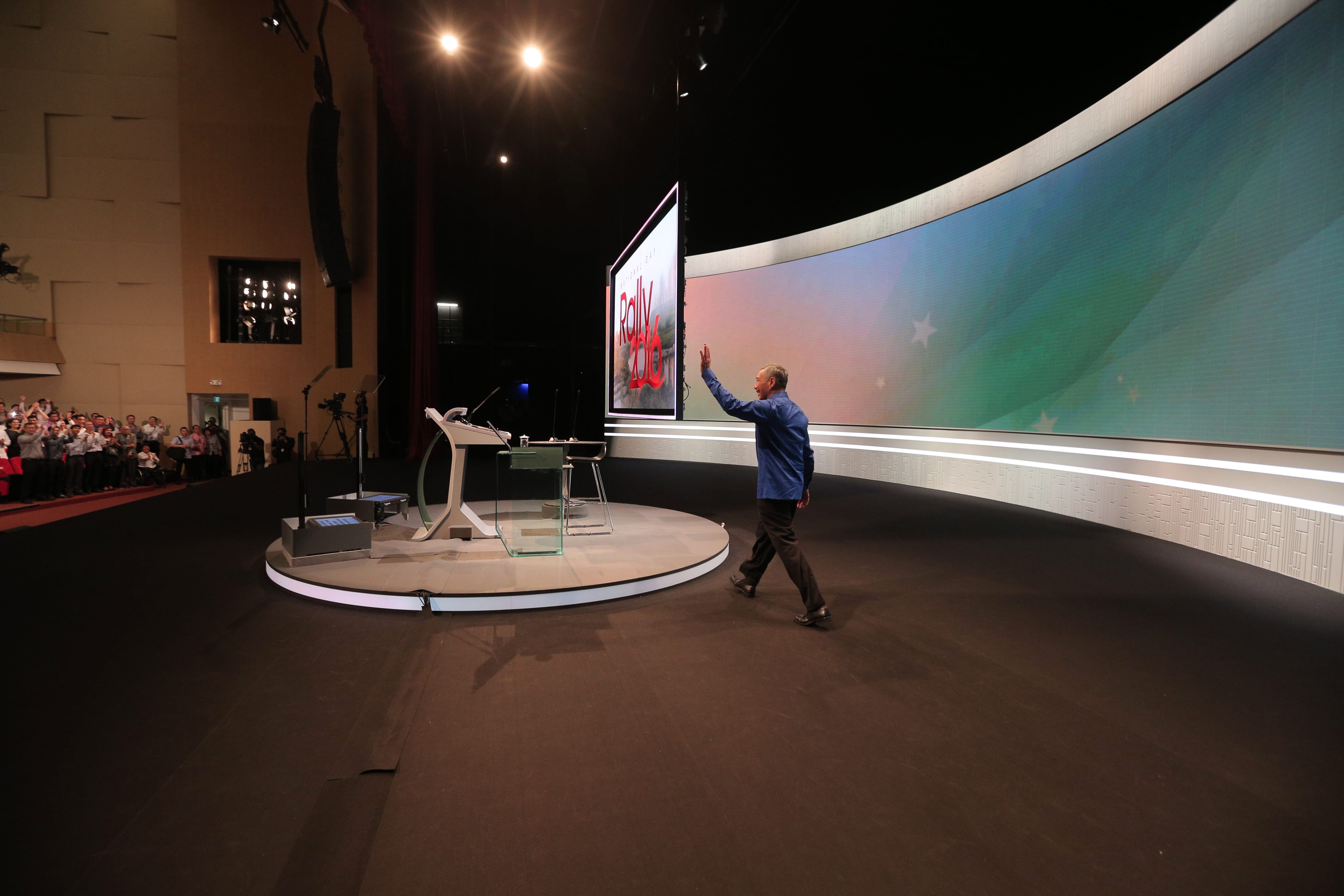 Man in blue shirt waves to screen onstage with Rally 2016 graphic and crowd background.