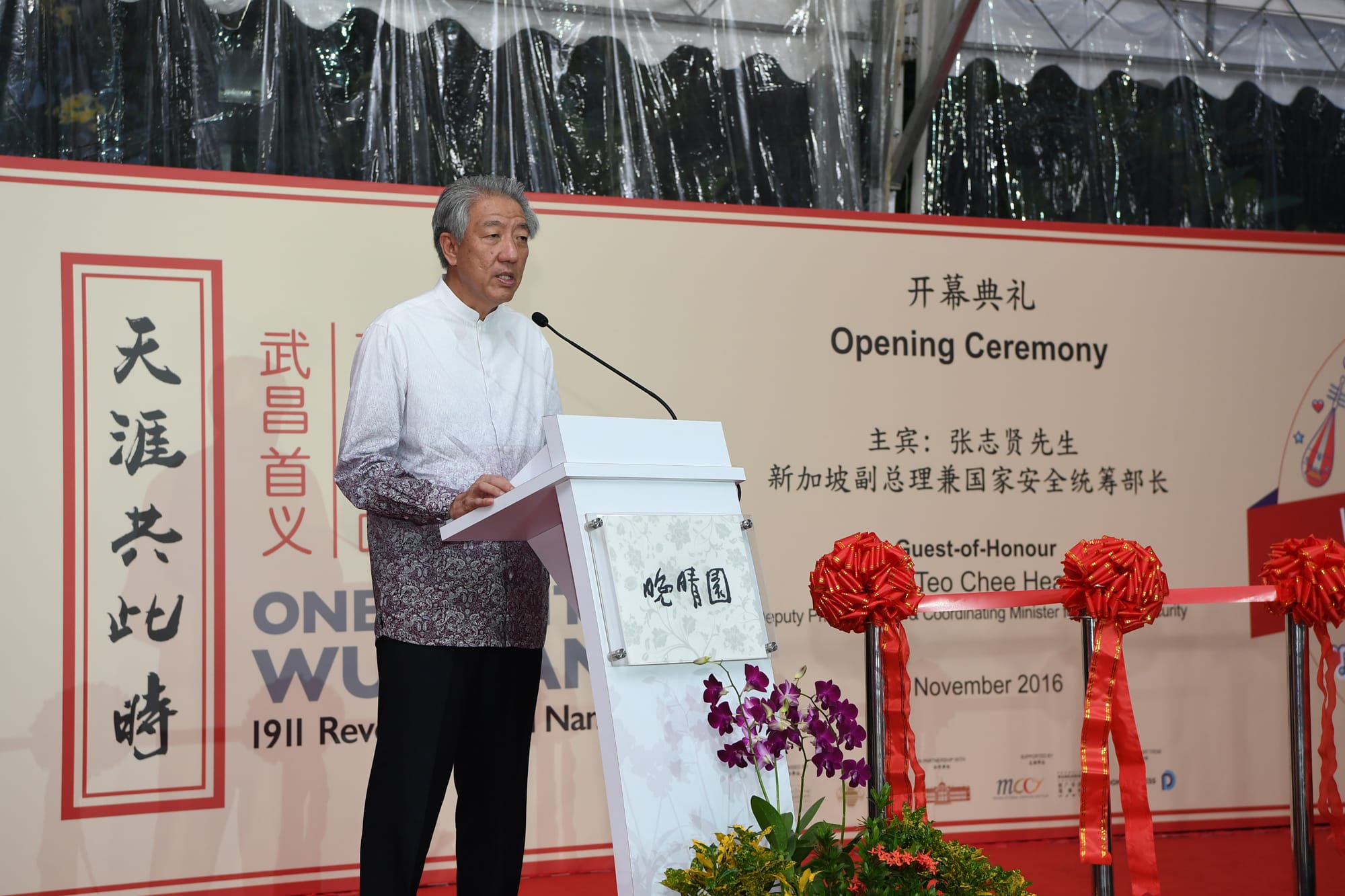 Man at podium with mic, speaking at an opening ceremony in front of a sign with Chinese characters.