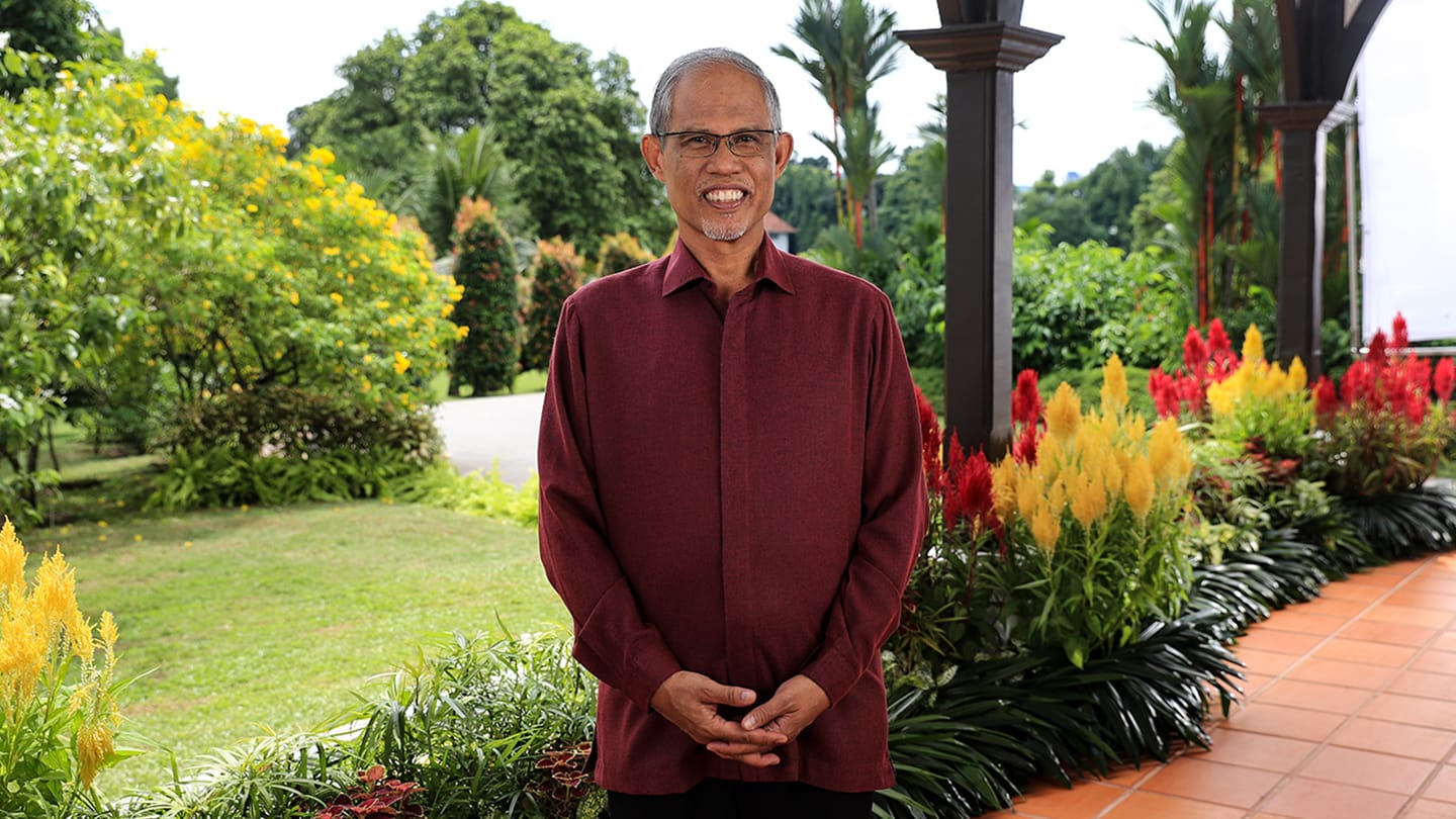 Man in glasses, maroon shirt, standing with hands clasped in a garden setting.