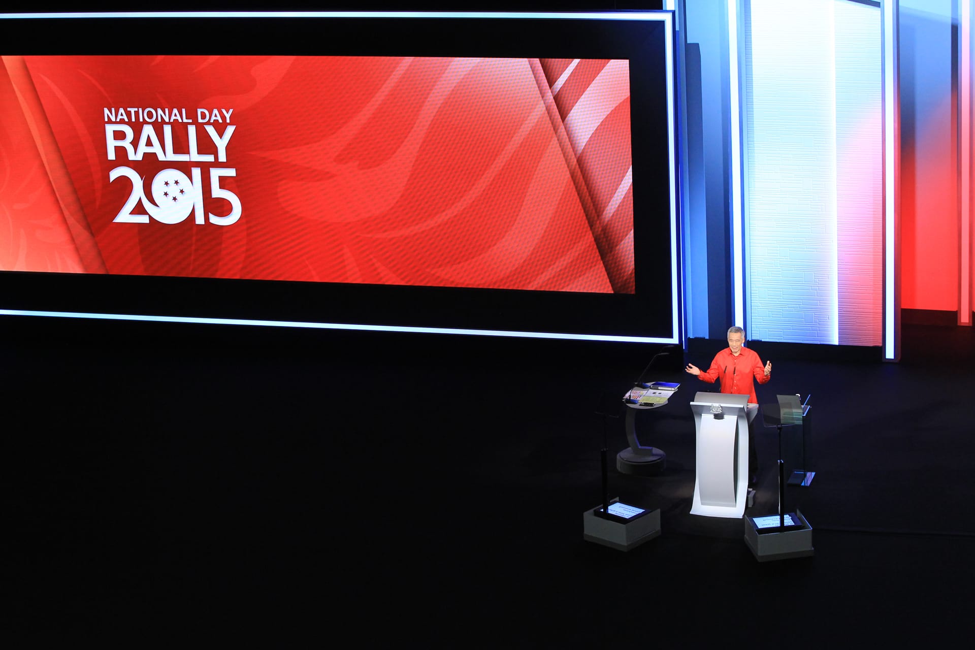 Man in red shirt at podium. Screen reads "National Day Rally 2015." Red, white, and blue background.