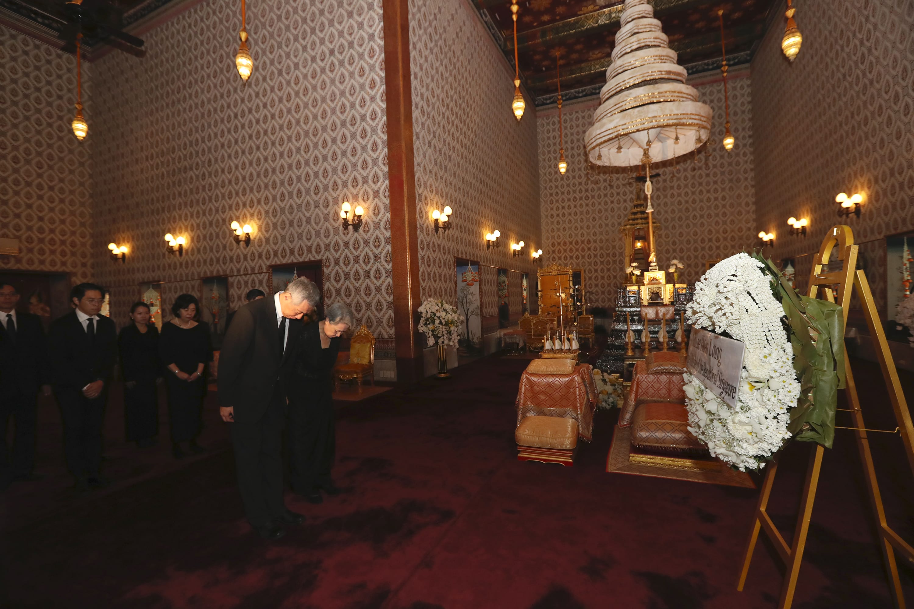 People in black attire bowing inside a building with ornate decor and floral wreaths.