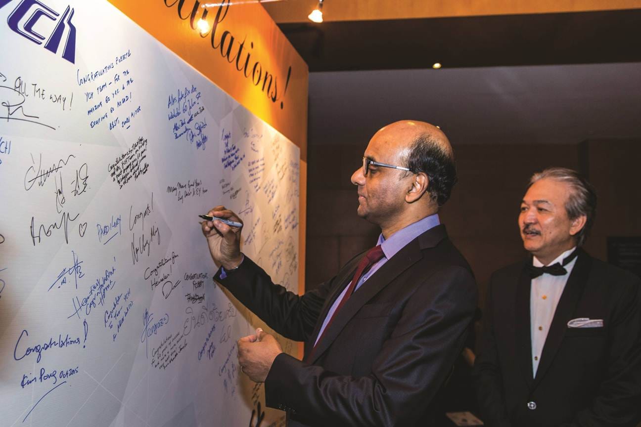Man signing a white wall with "Congratulations!" messages. Another man in a tuxedo watches.