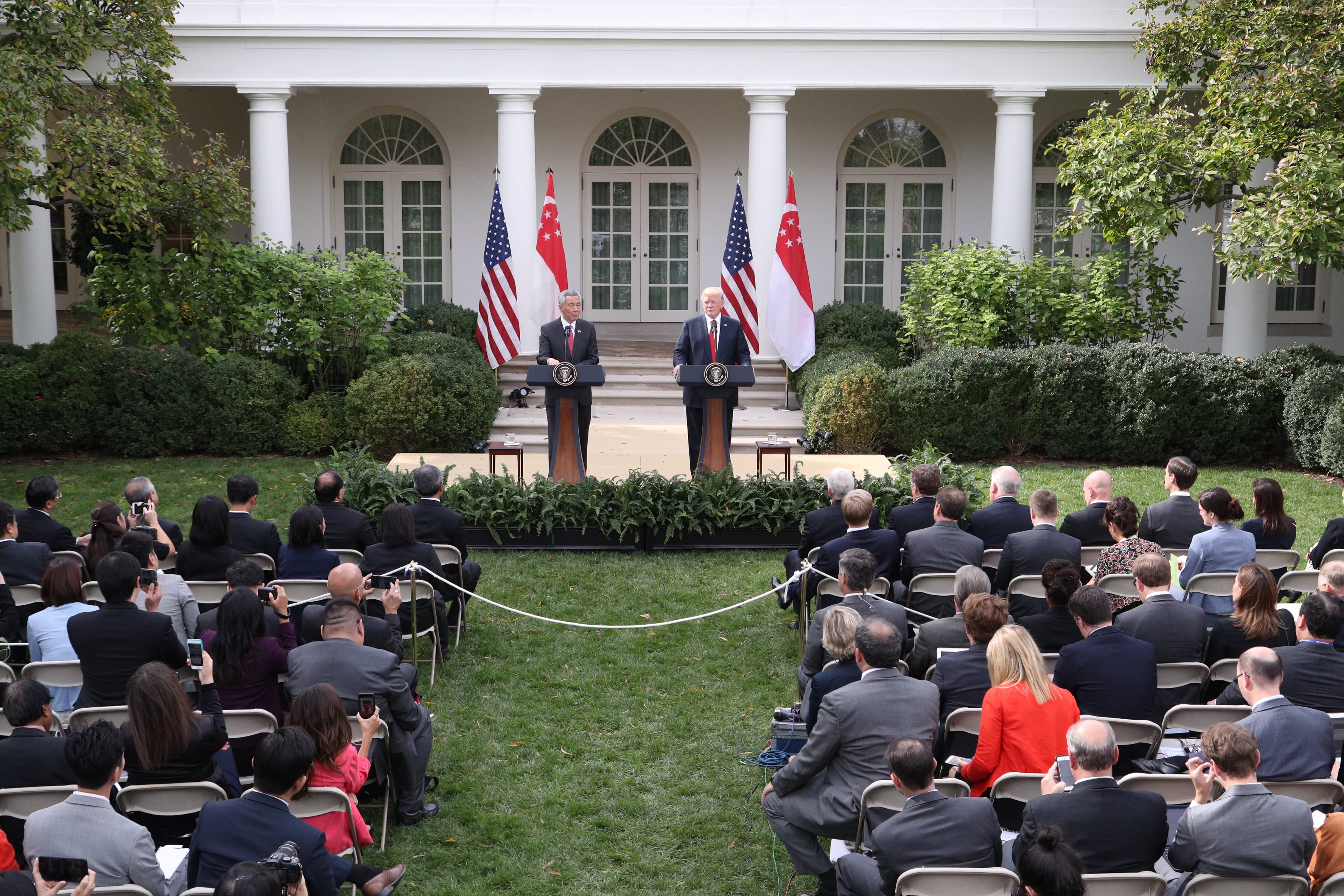 Trump and Lee Hsien Loong at podiums on lawn before crowd. US and Singapore flags behind.