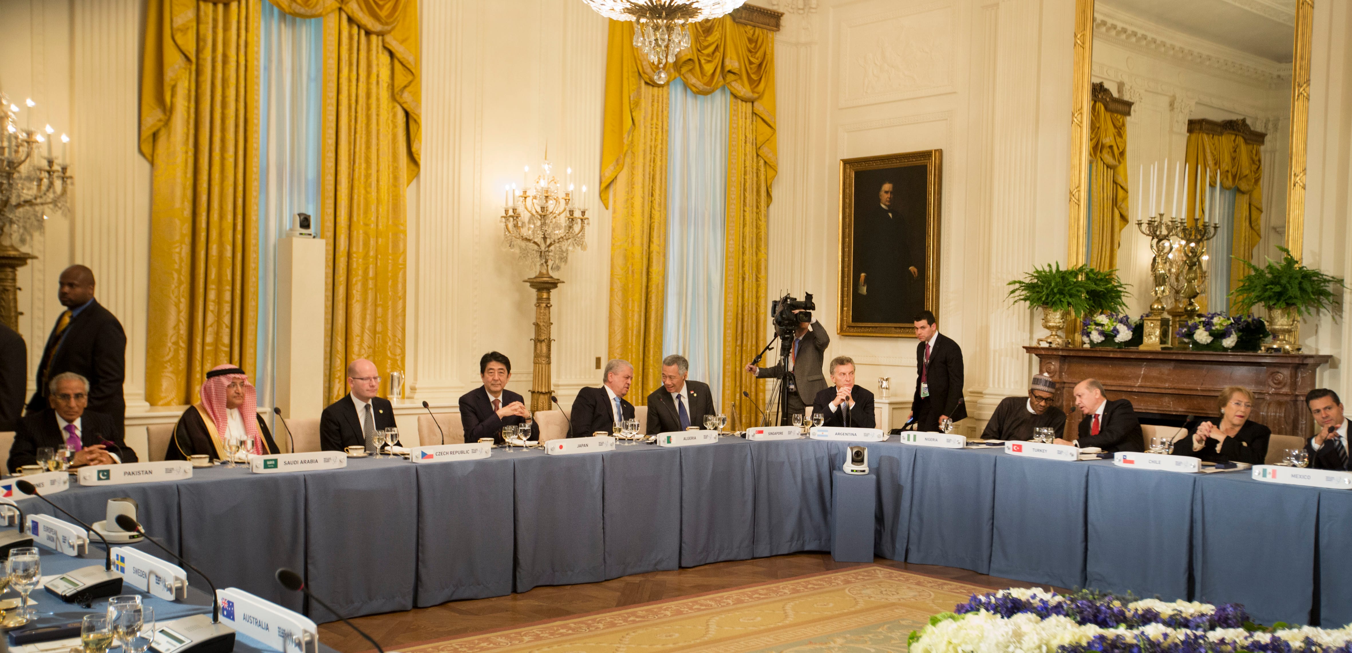 Leaders seated at long blue-clothed table with country nameplates in a grand room.