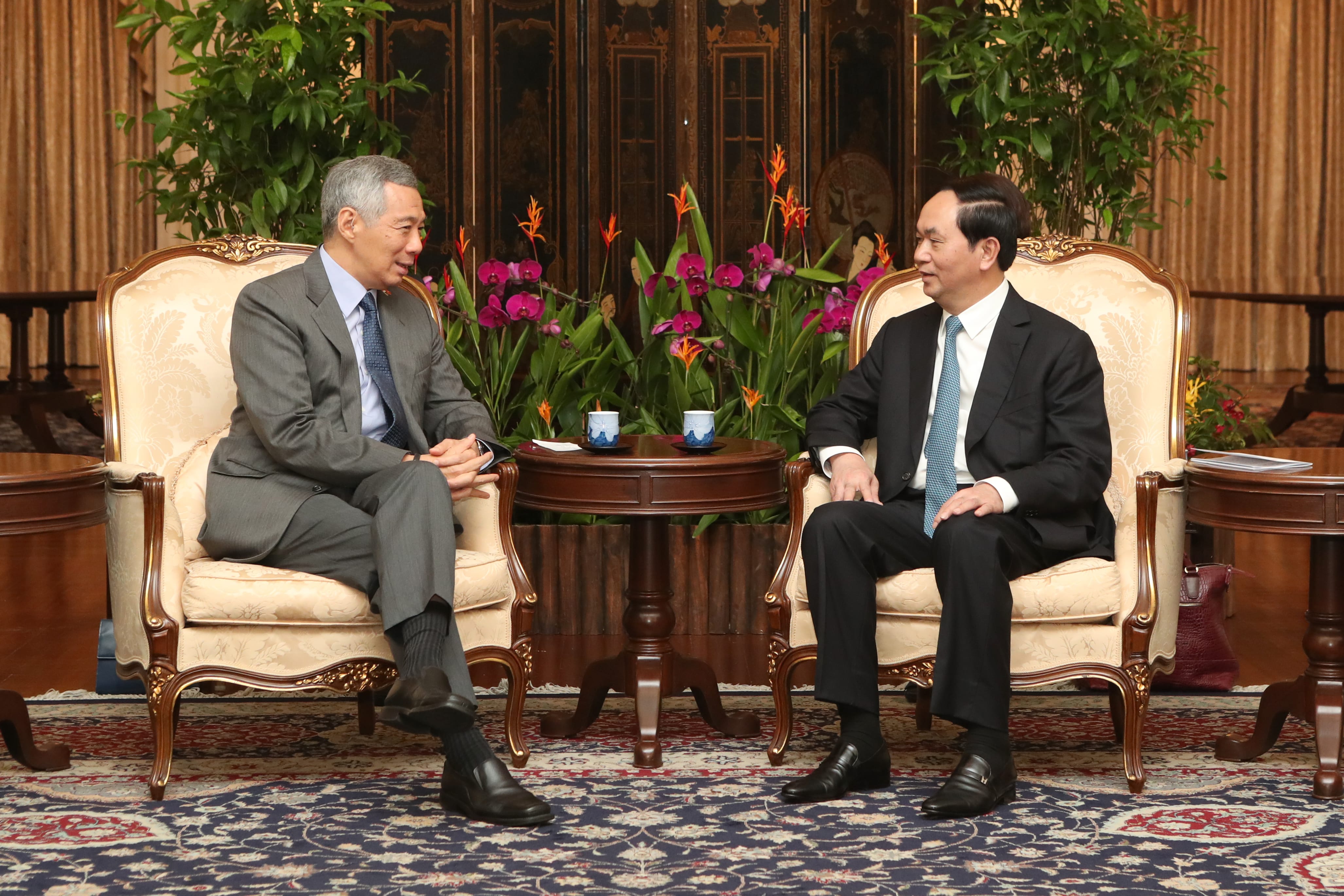 Two men in suits sit facing each other in ornate chairs with flower displays in the background.
