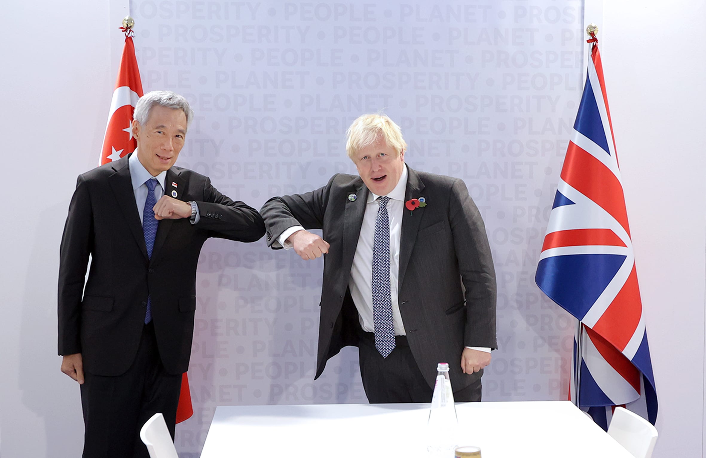 Two men, Lee Hsien Loong and Boris Johnson, bump elbows in front of flags of Singapore and the UK.