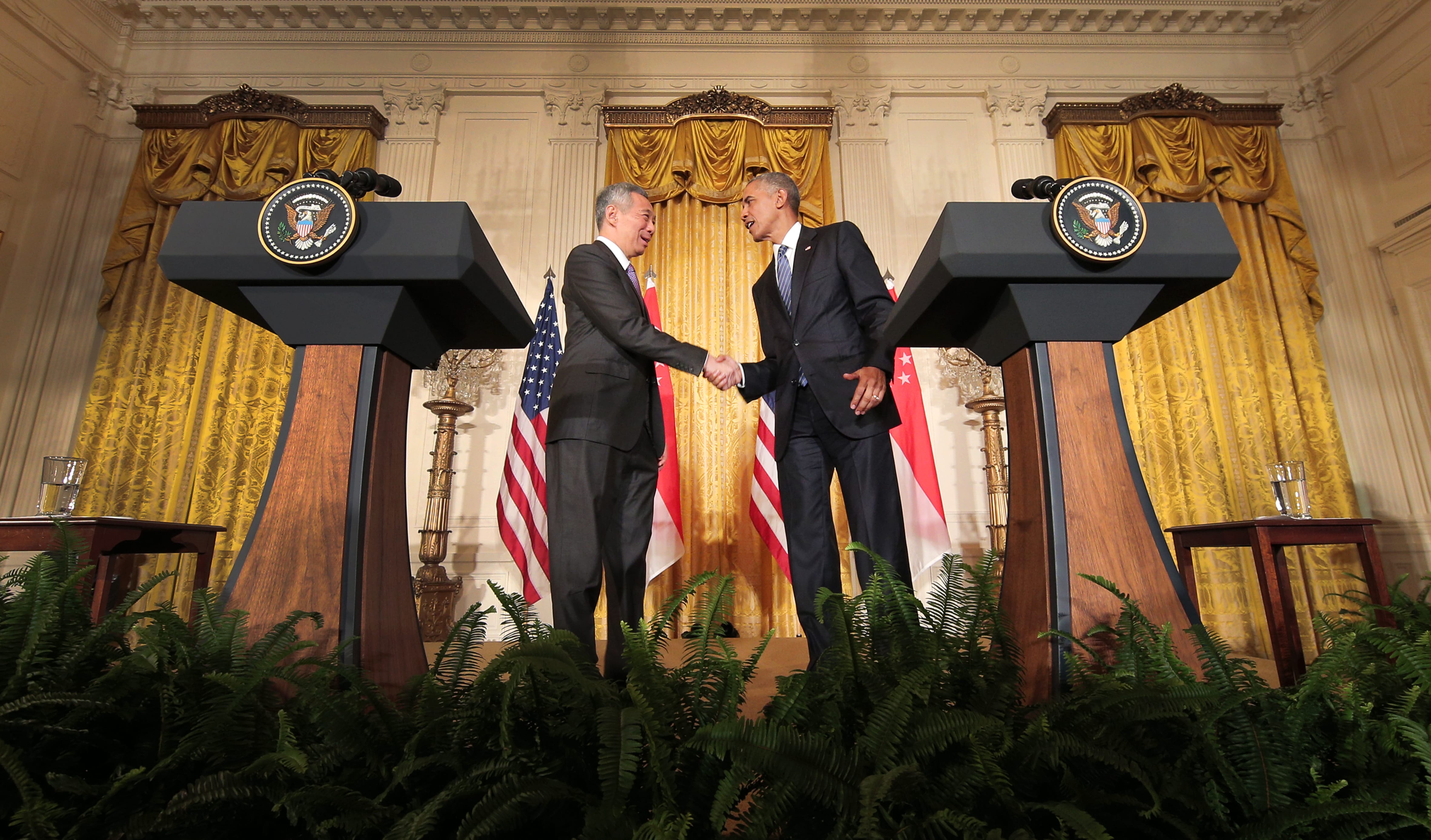 Lee Hsien Loong and Barack Obama in suits shake hands near podiums with US seals.