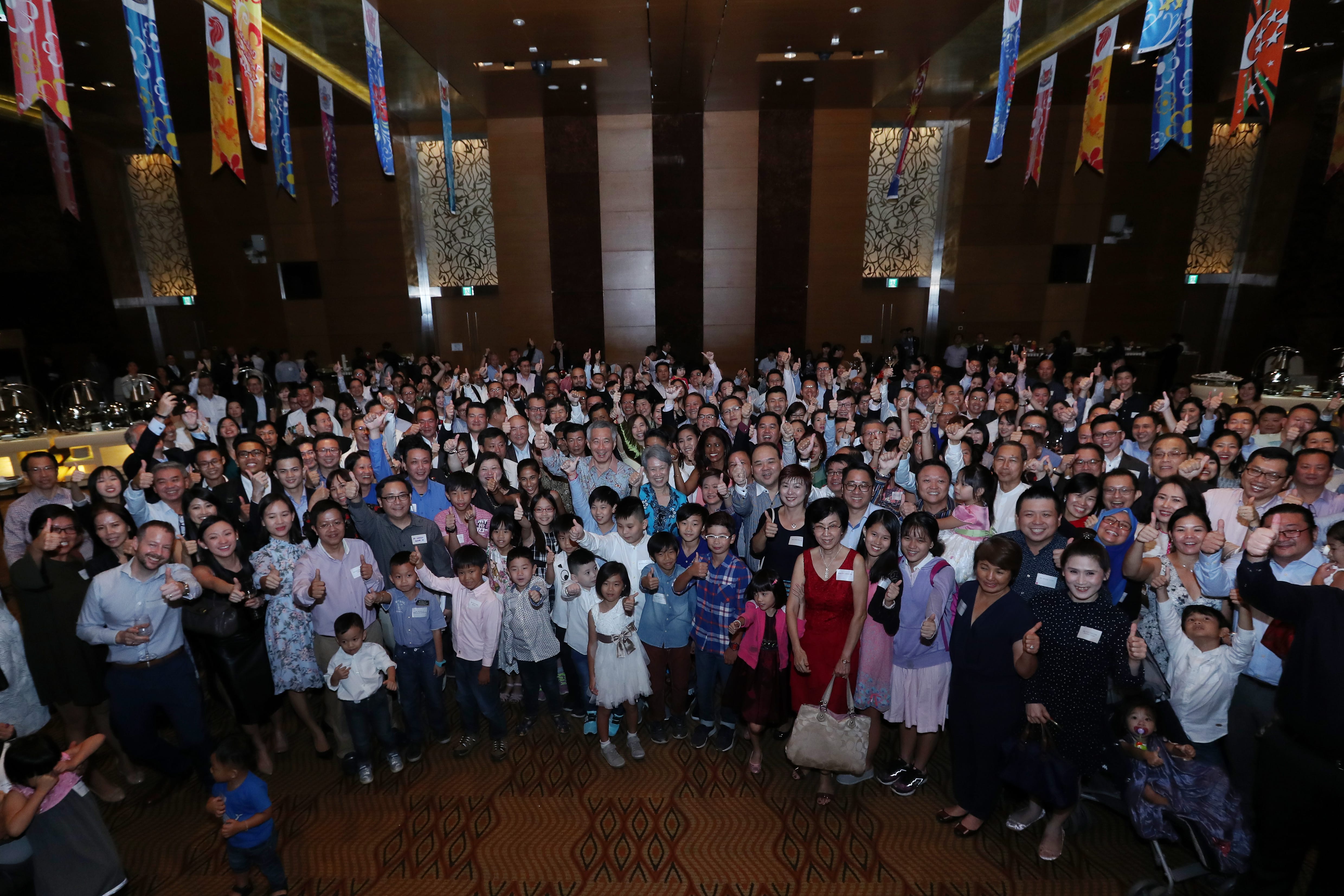Large group giving thumbs up in decorated hall with Lee Hsien Loong visible.
