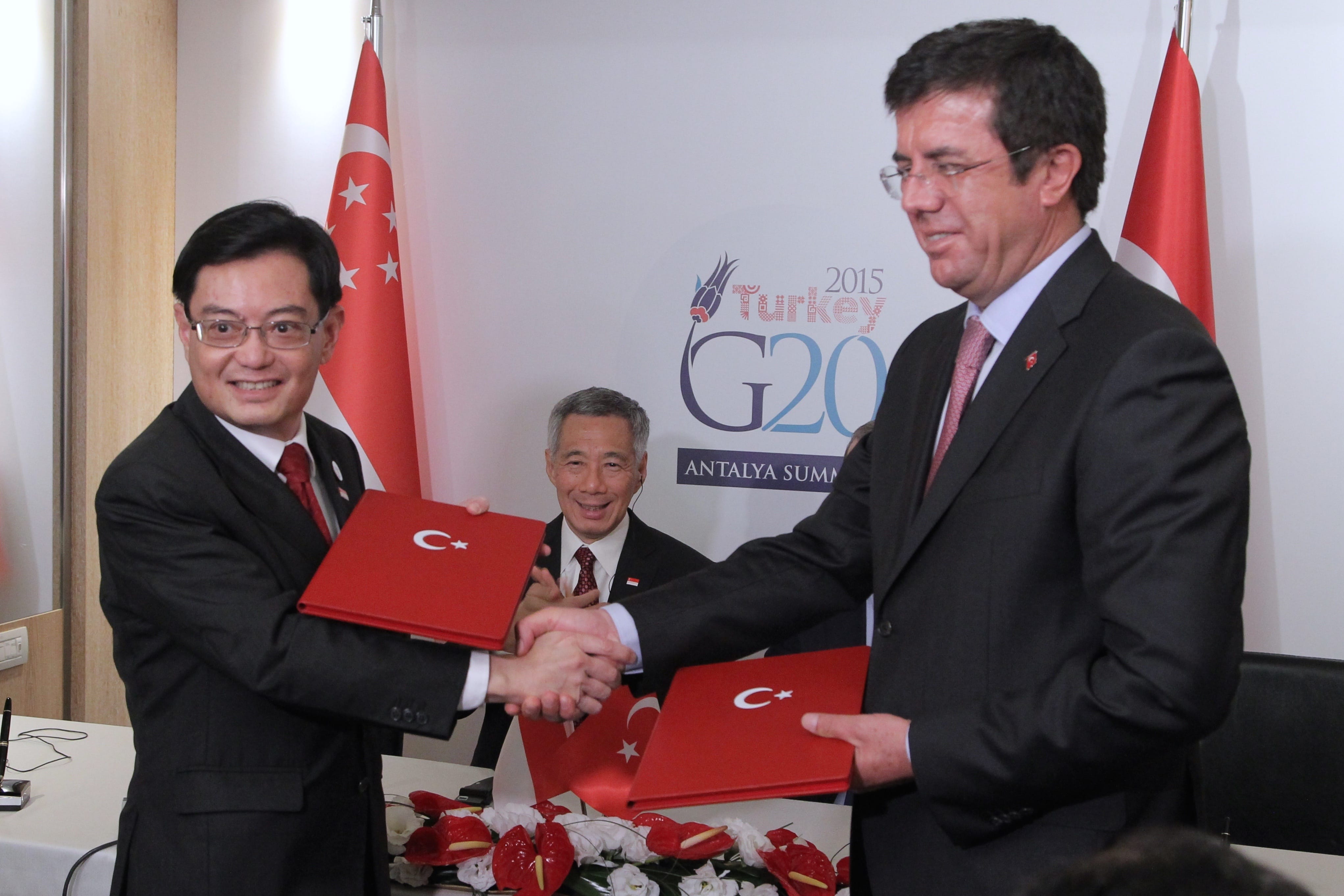 Two men in suits shake hands, holding red folders with Turkish flag. Singapore and Turkey flags behind them. G20 summit logo in background.