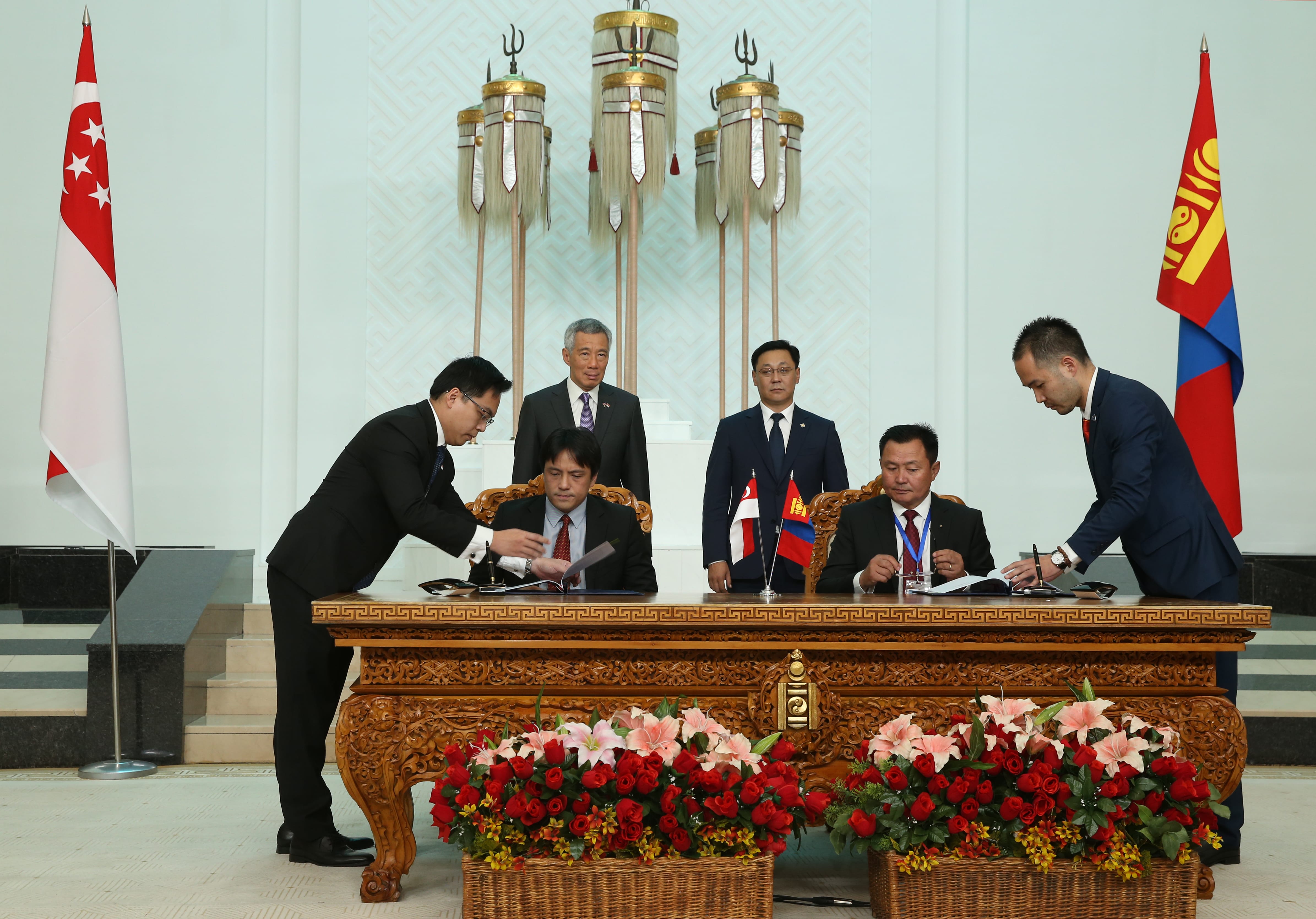 Two men sign documents at a decorated table with Singapore and Mongolia flags. Lee Hsien Loong stands behind.