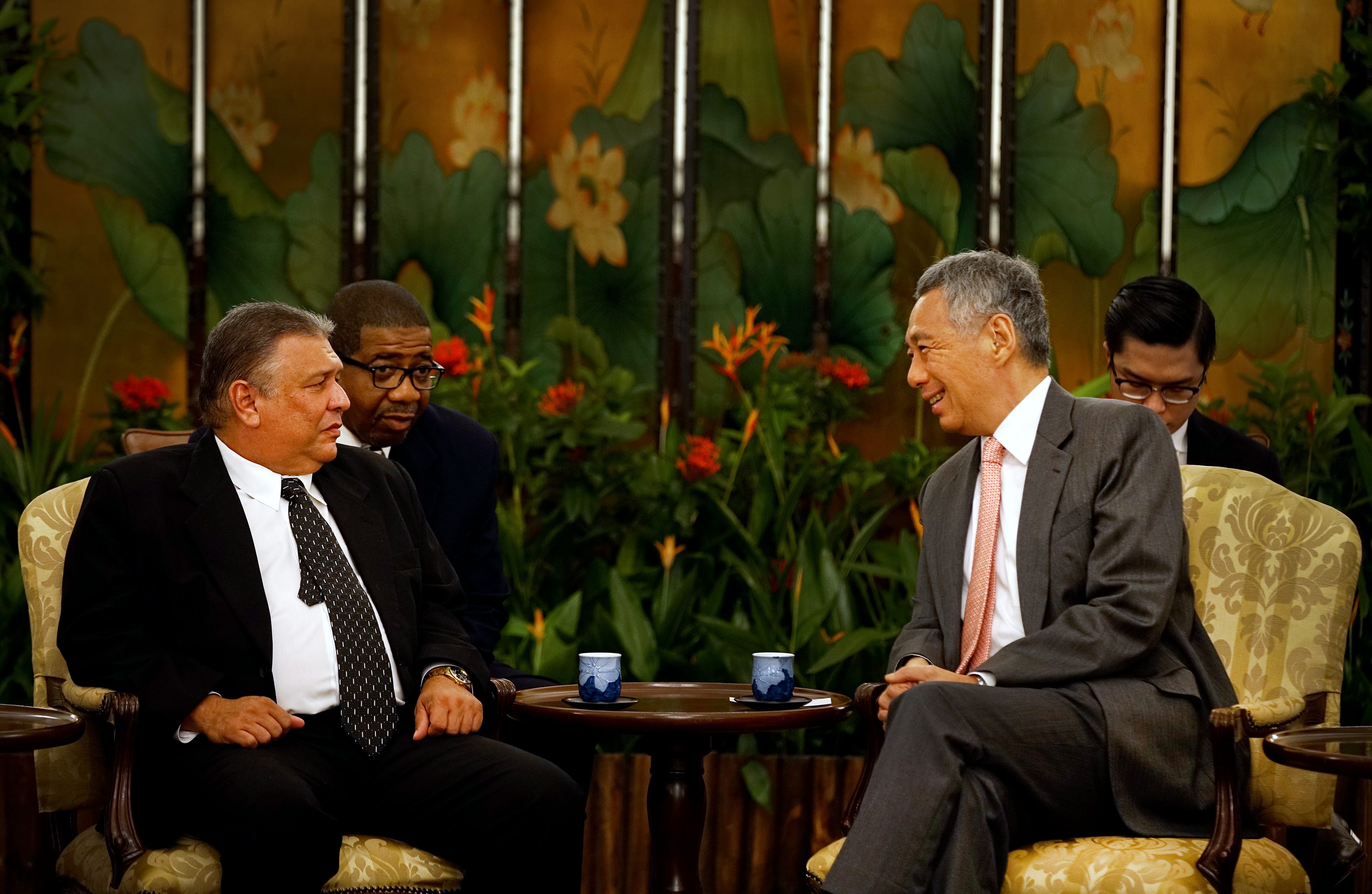 Two men in suits sit facing each other, with two men behind them. All are in an ornate room.