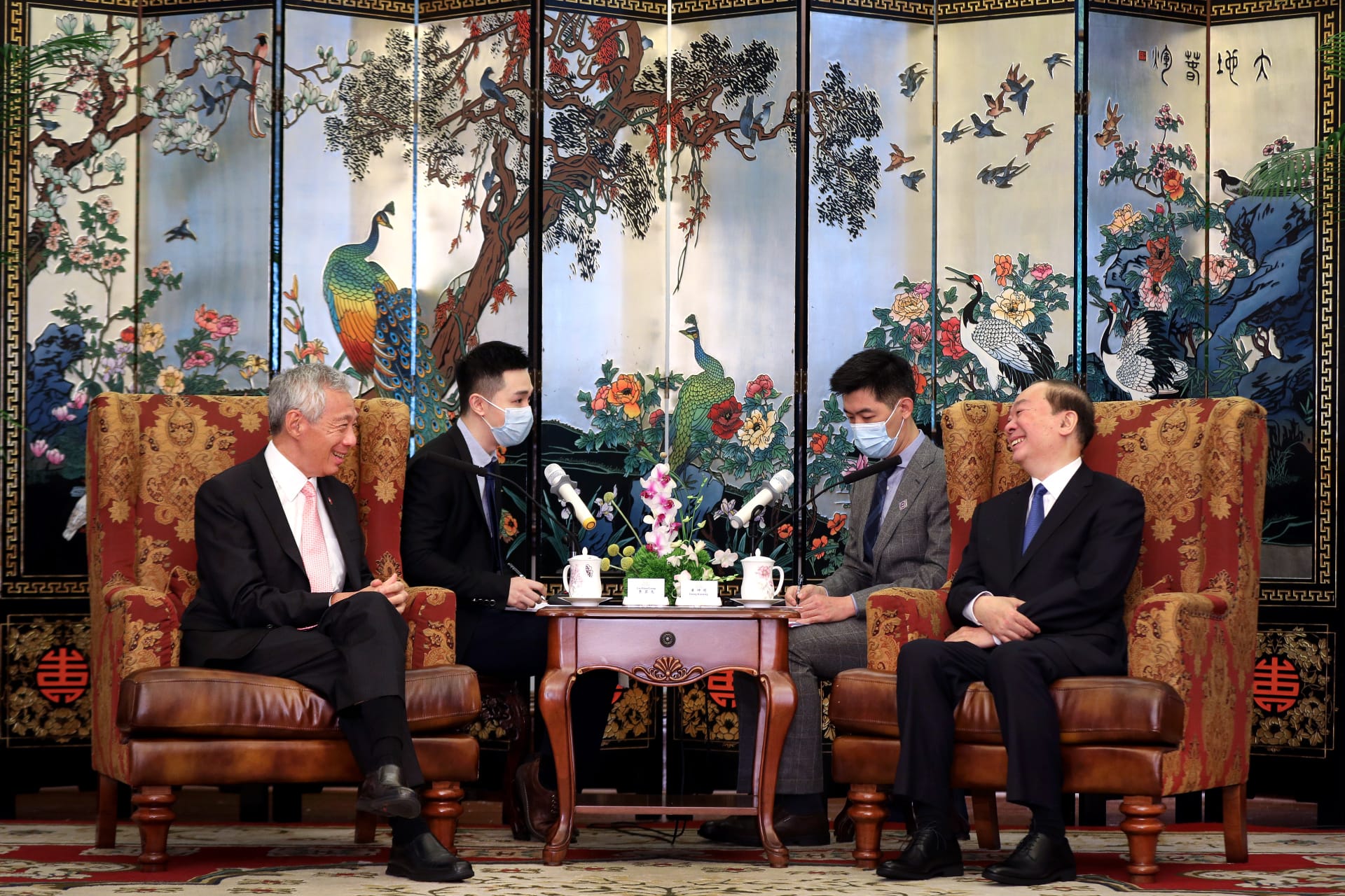 Lee Hsien Loong and another man sit facing each other wearing masks. Behind them is a painted folding screen.