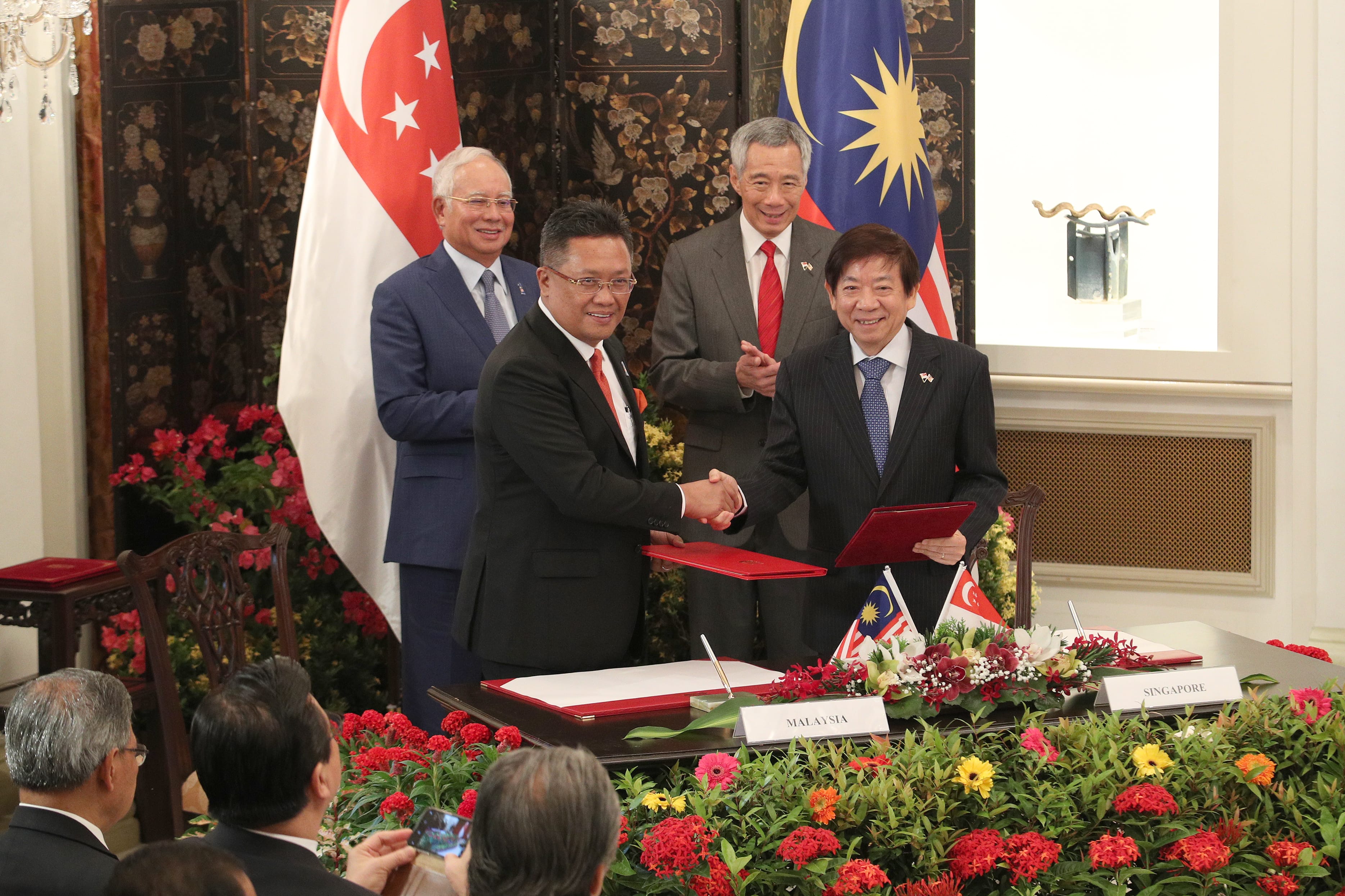 Four men in suits, including Lee Hsien Loong, shake hands over a table with Singapore and Malaysia flags.