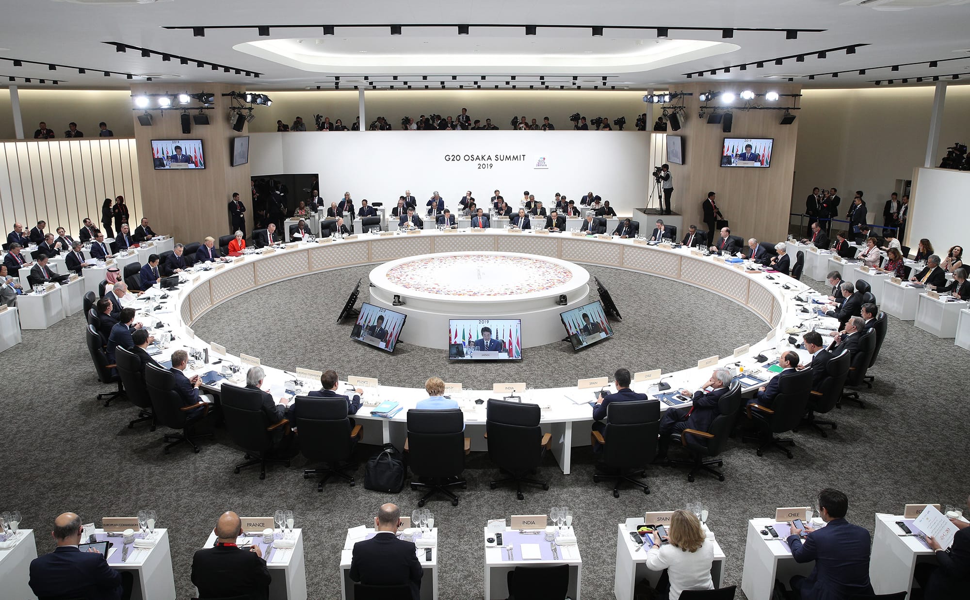 G20 Osaka Summit 2019, leaders seated in a circle around a raised platform with screens. Attendees in gallery.