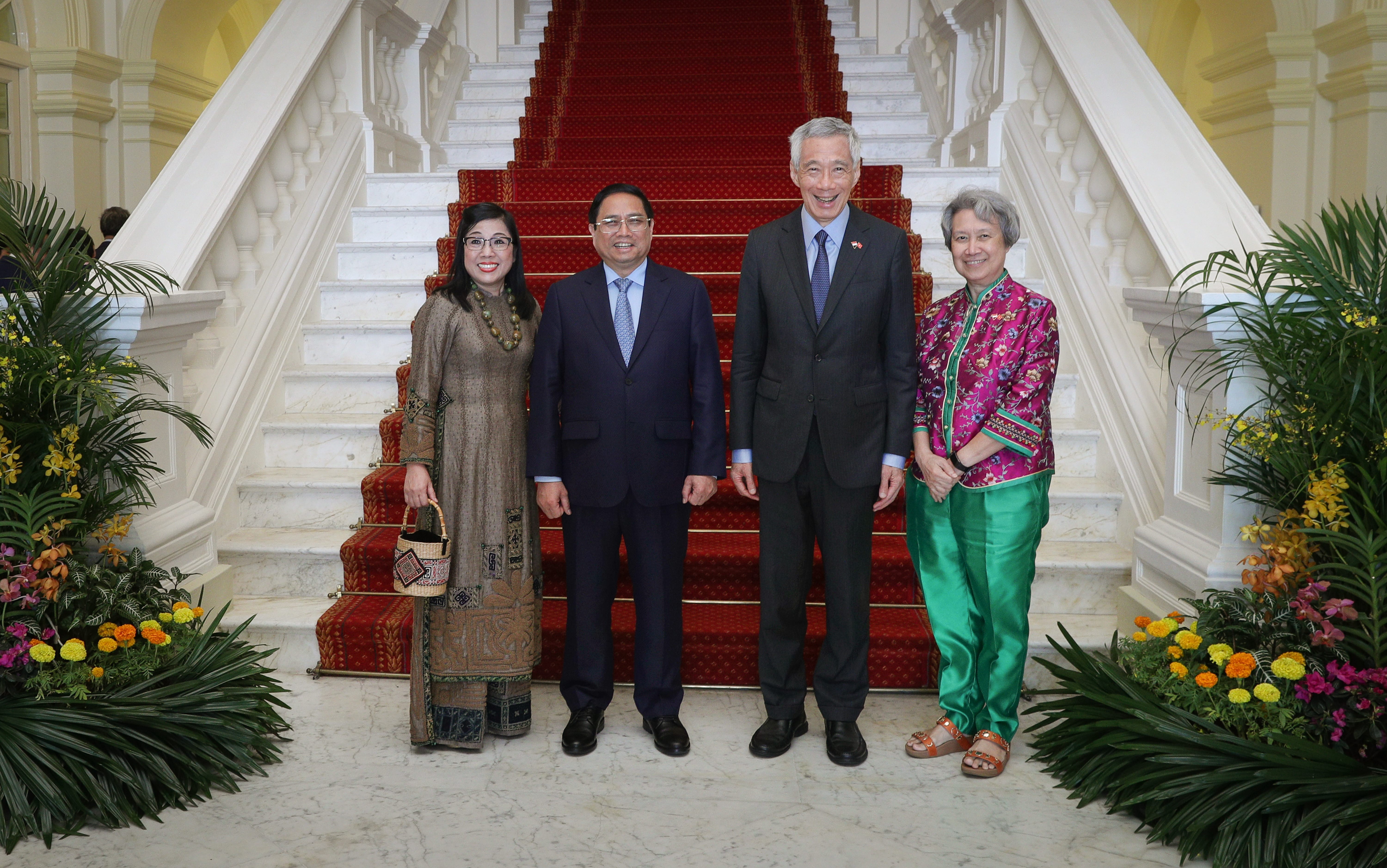 Four people pose on a white staircase with a red runner, surrounded by greenery and flowers.
