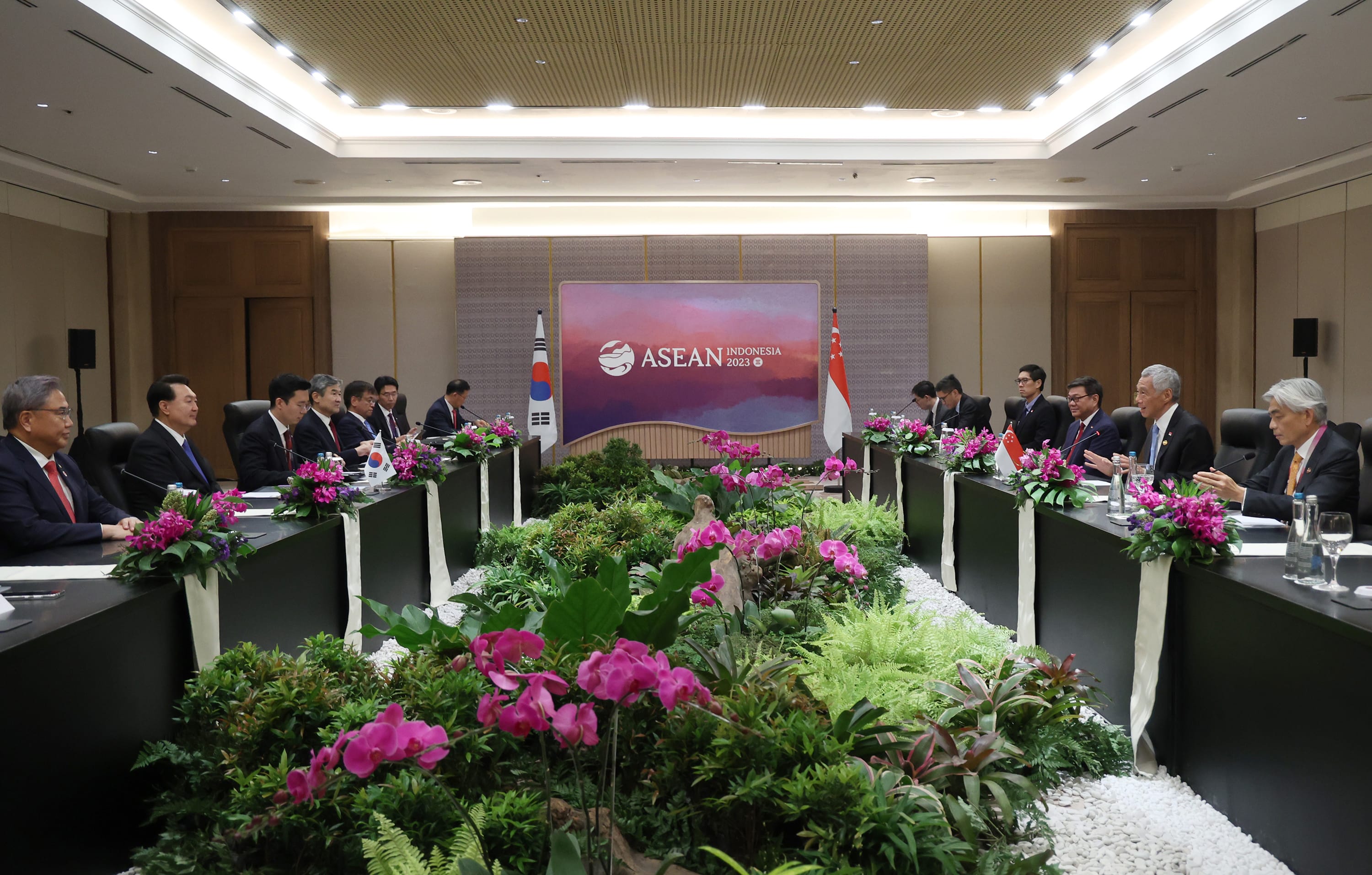 Meeting room with leaders at long tables, flags of South Korea and Singapore, ASEAN Indonesia 2023 backdrop, floral arrangements.