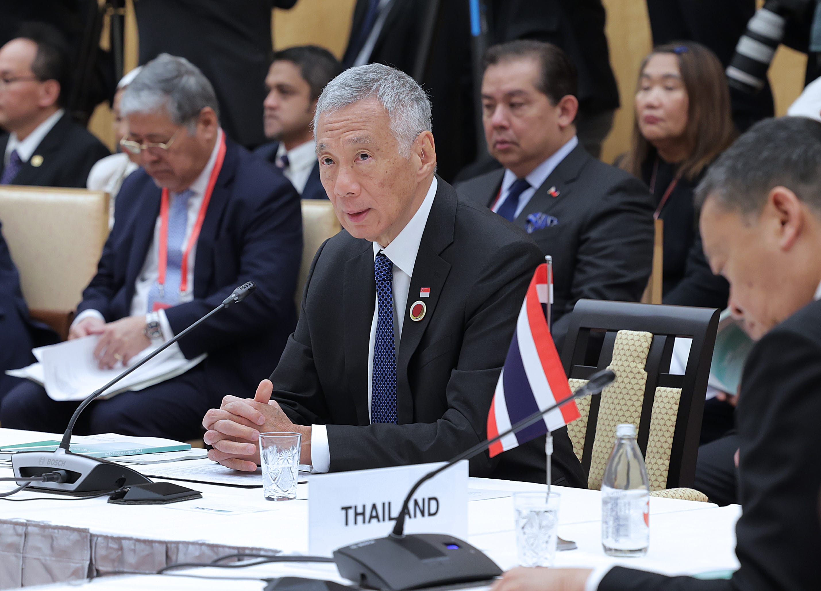 Lee Hsien Loong at a conference table, wearing a suit with a Singapore flag pin. "Thailand" sign is visible.