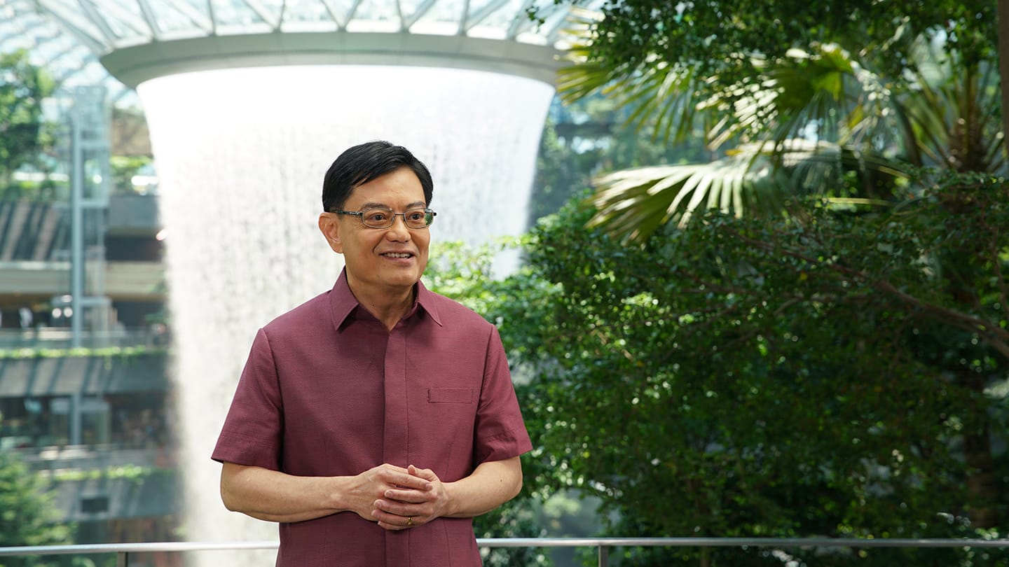 Lawrence Wong wearing glasses, in a maroon shirt with Jewel Changi Airport's Rain Vortex behind him.
