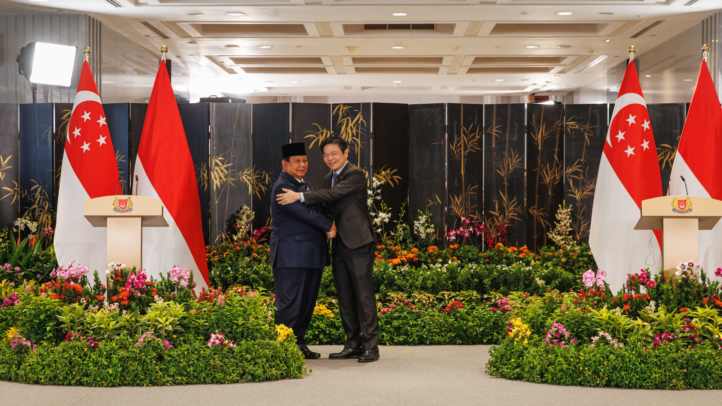 Two men in suits shake hands before a floral backdrop and Singaporean/Indonesian flags.